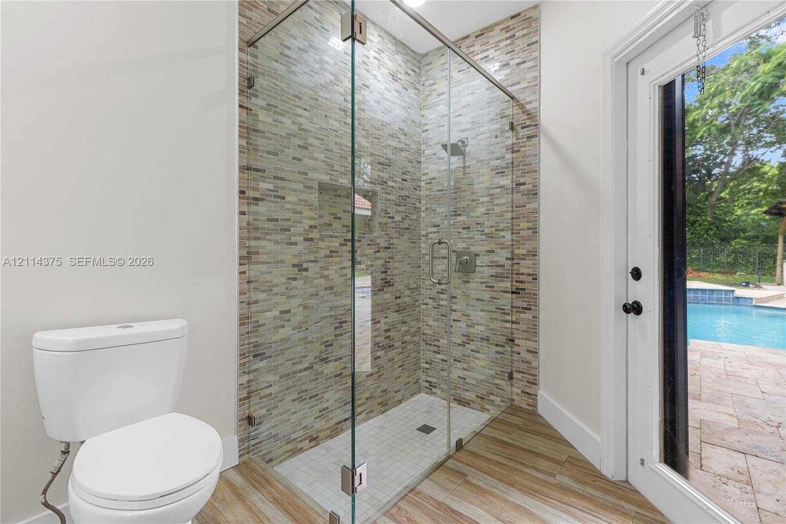 1206 S Manor Drive S, Weston, Weston, FL 33326 Walk-in shower featuring a frameless glass enclosure, recessed wall niche, and textured multi-tonal tile surround, complemented by wood-finish flooring and direct external access