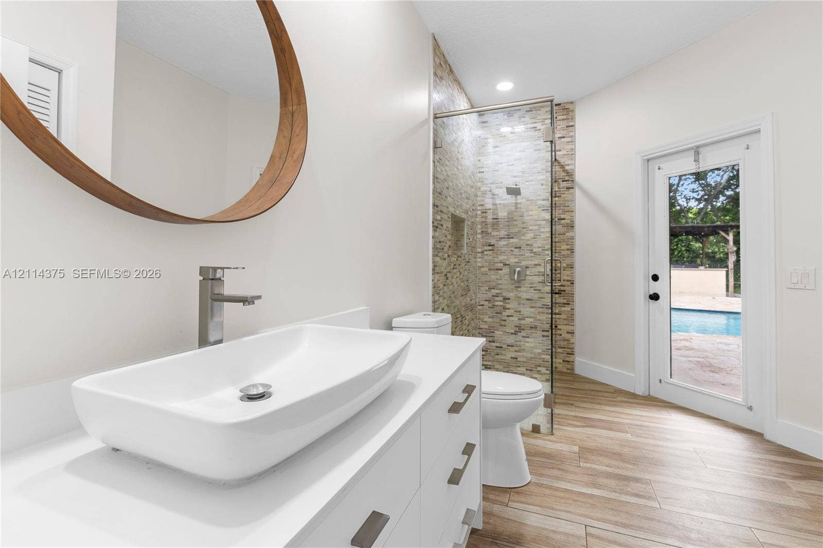 1206 S Manor Drive S, Weston, Weston, FL 33326 Bathroom featuring a rectangular vessel sink with a chrome faucet, wood-framed circular mirror, walk-in shower with stone-tile surround, wood-finish flooring, and a glass-paneled exterior door