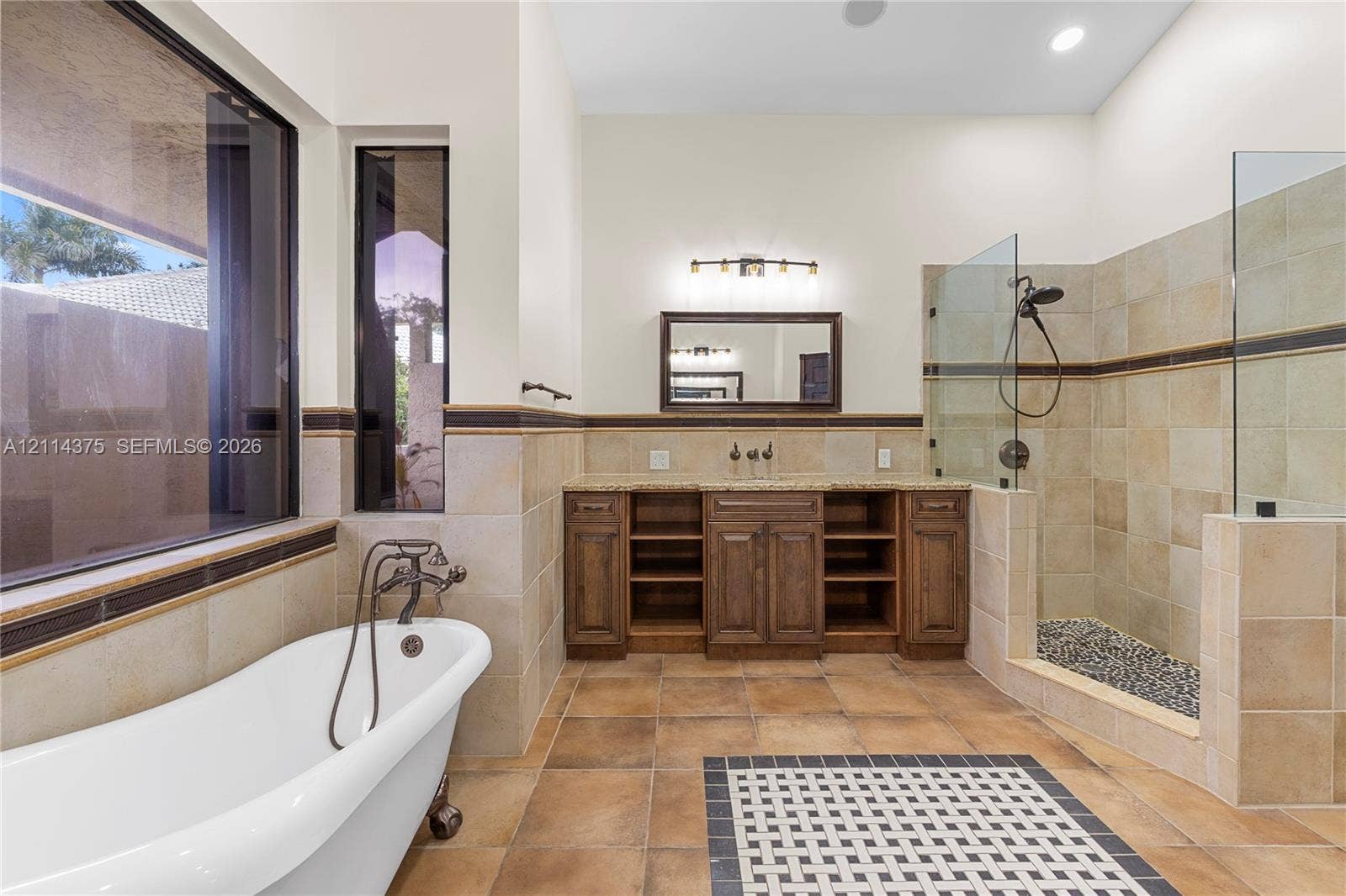1206 S Manor Drive S, Weston, Weston, FL 33326 Freestanding soaking tub with dark metal faucet, dual-sink wooden vanity with stone countertop, tiled walk-in shower with glass enclosure, and patterned tile floor inlay