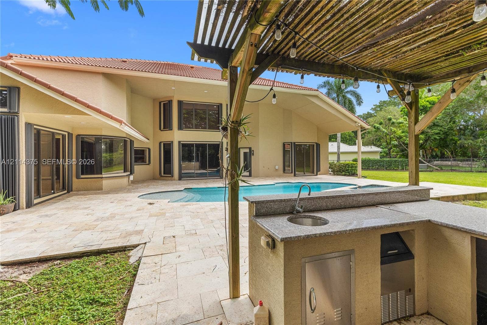 1206 S Manor Drive S, Weston, Weston, FL 33326 Outdoor kitchen featuring stone-finish counters, integrated sink, and stainless-steel cabinetry under a wooden pergola, situated on a stone-paver patio with a custom pool