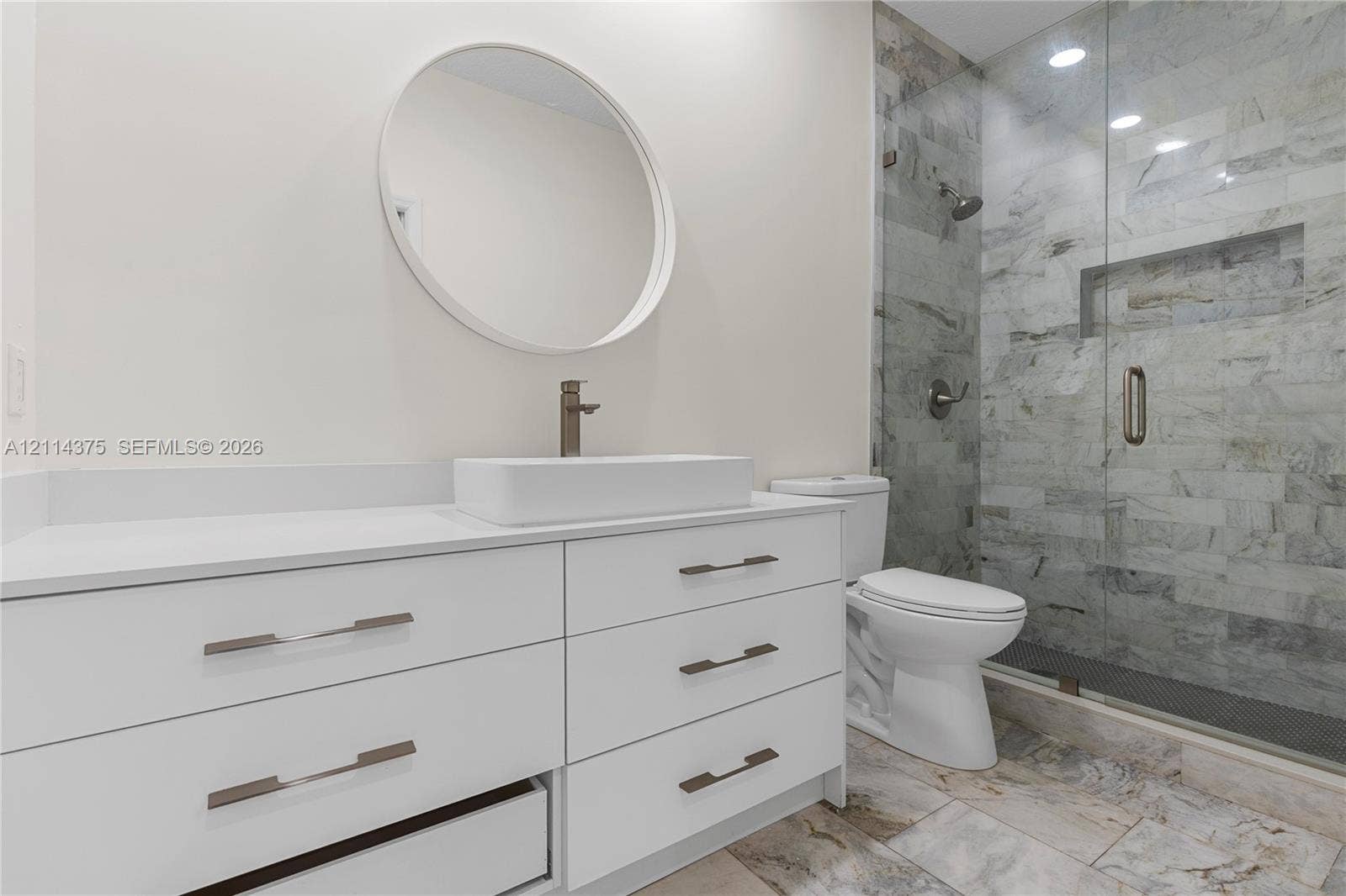 1206 S Manor Drive S, Weston, Weston, FL 33326 White cabinetry vanity with rectangular vessel sink, frameless glass shower enclosure with stone-tile surround, built-in shower niche, and round wall-mounted mirror