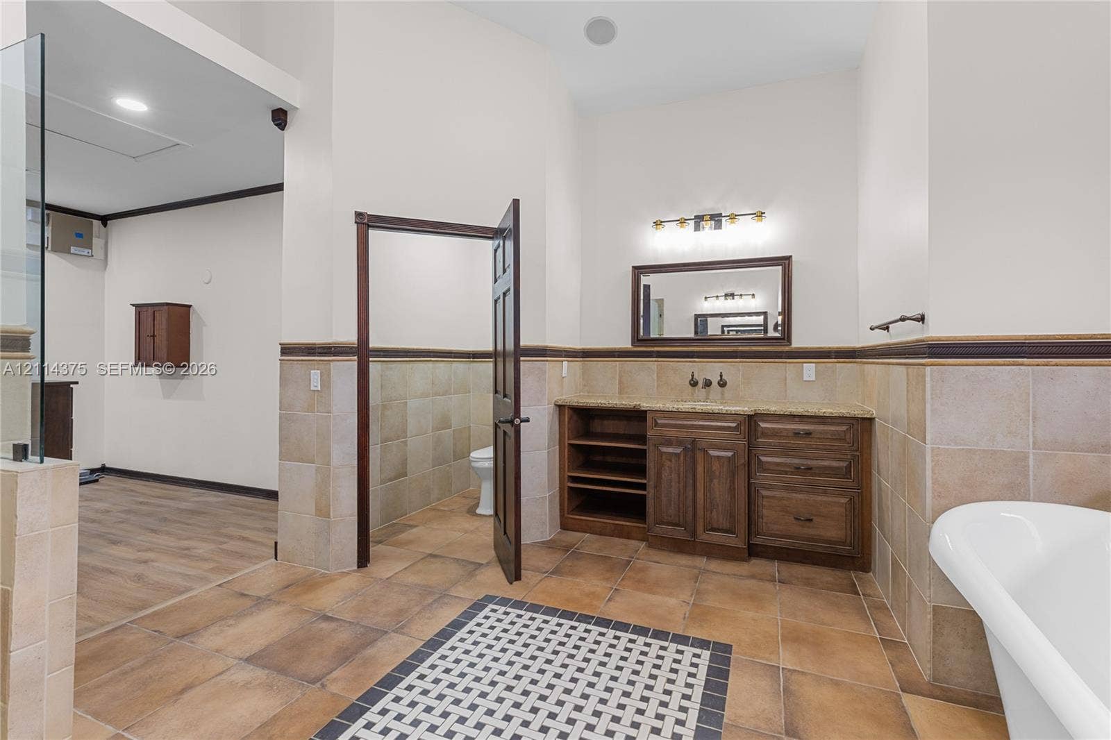 1206 S Manor Drive S, Weston, Weston, FL 33326 Bathroom featuring a wood-tone vanity with stone countertop, tile wainscoting, a wall-mounted framed mirror, a soaking tub, and a decorative tile inlay floor