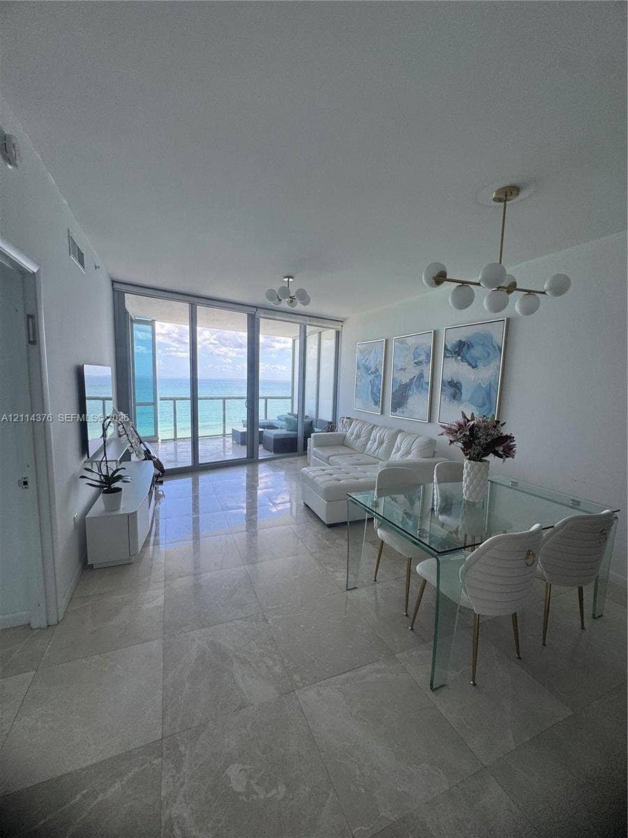 Open-concept living space featuring floor-to-ceiling glass panels, large-format tile flooring, multi-globe brass-finish chandeliers, and a private balcony with ocean frontage