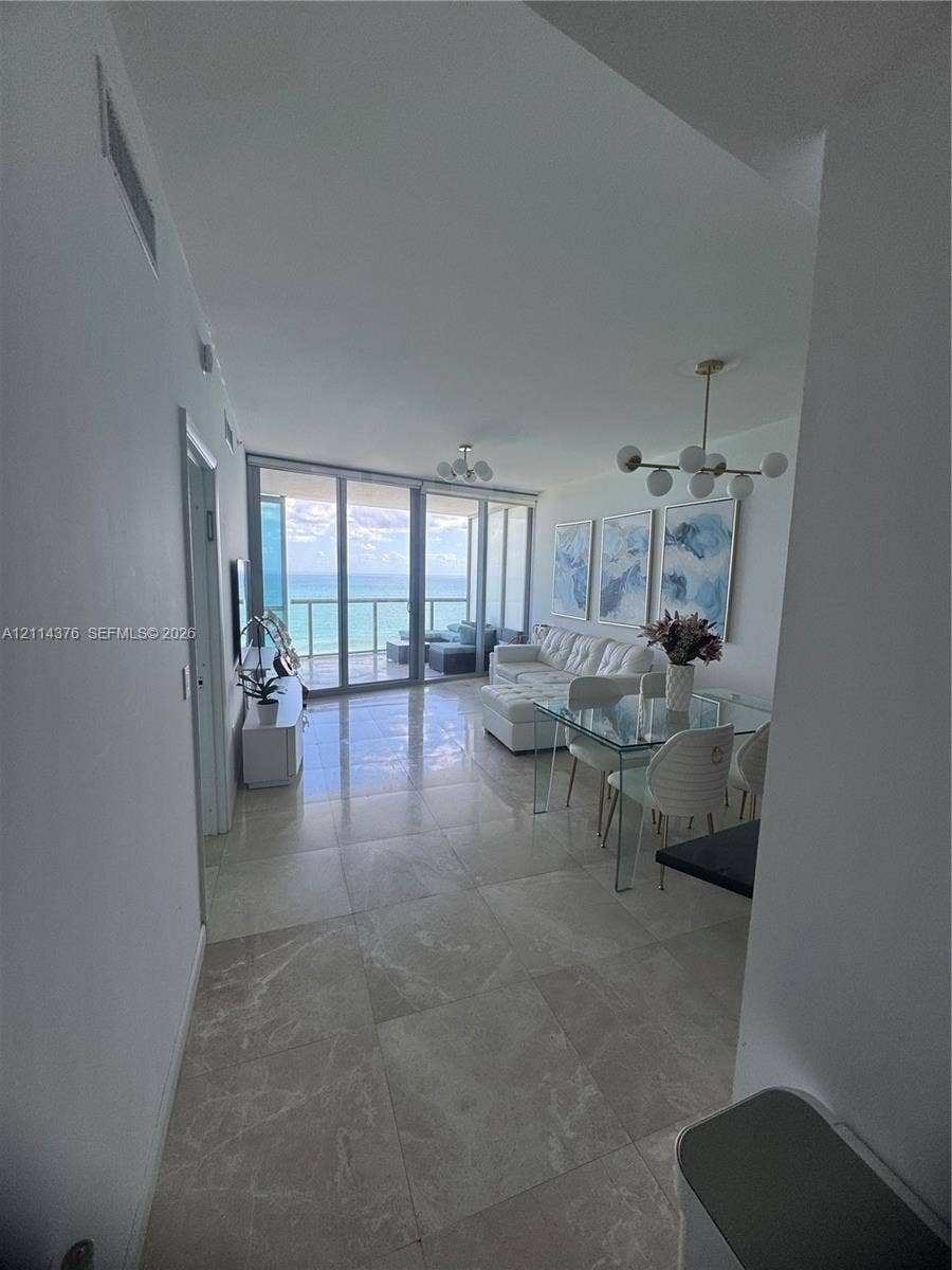 Large-format tile flooring, multi-globe metallic chandelier, floor-to-ceiling glass paneling, and an external balcony featuring a glass railing with an ocean backdrop