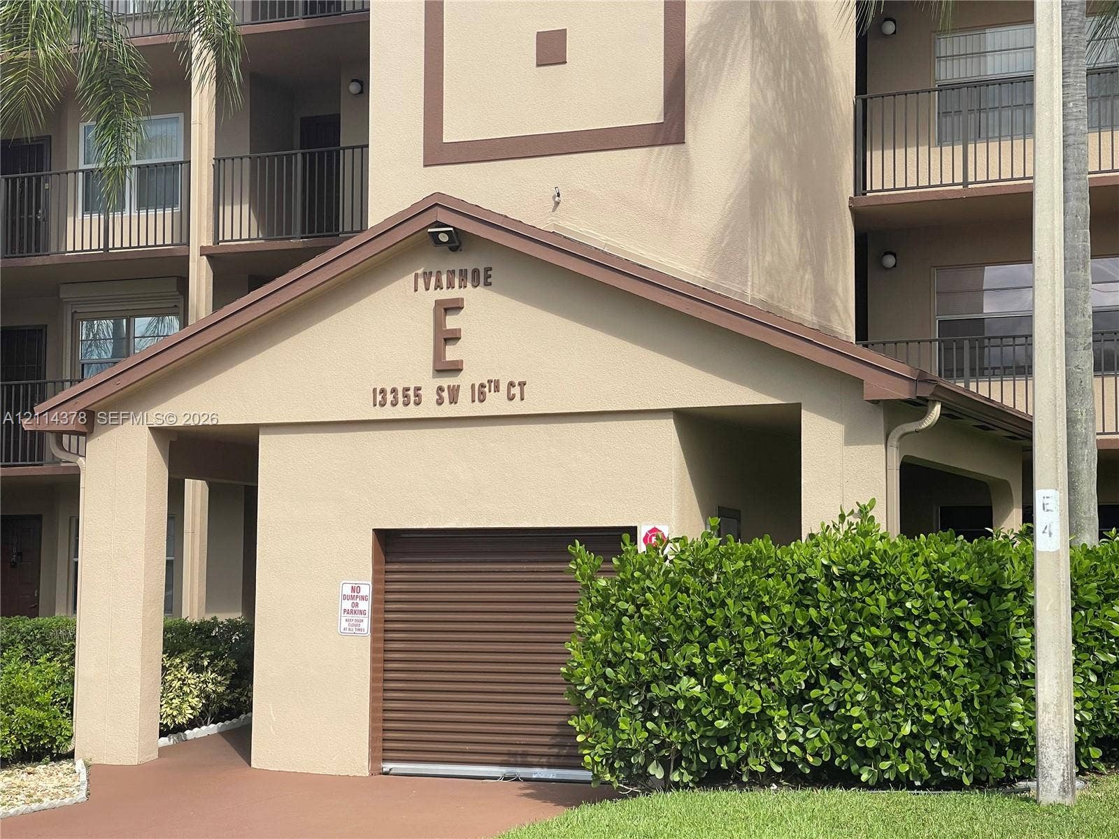 13355 SW 16th Court, #205E, Pembroke Pines, FL 33027 Multi-story residential building exterior featuring a stucco facade, covered entryway, private garage with a roll-up door, and iron balcony railings