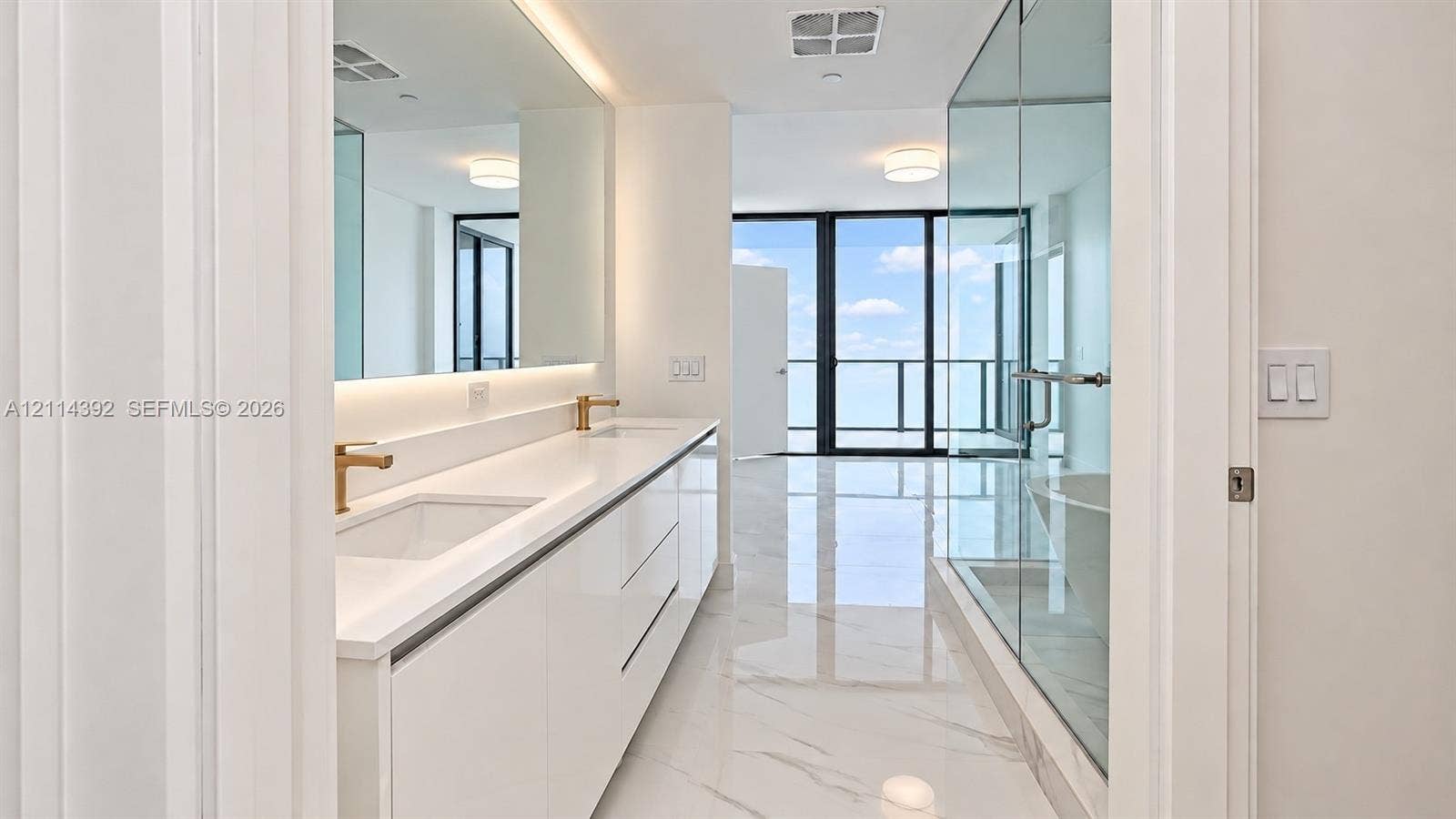Bathroom featuring white cabinetry, dual undermount sinks with brass-tone fixtures, wall-to-wall mirror, large-format tile flooring, and a glass-enclosed shower