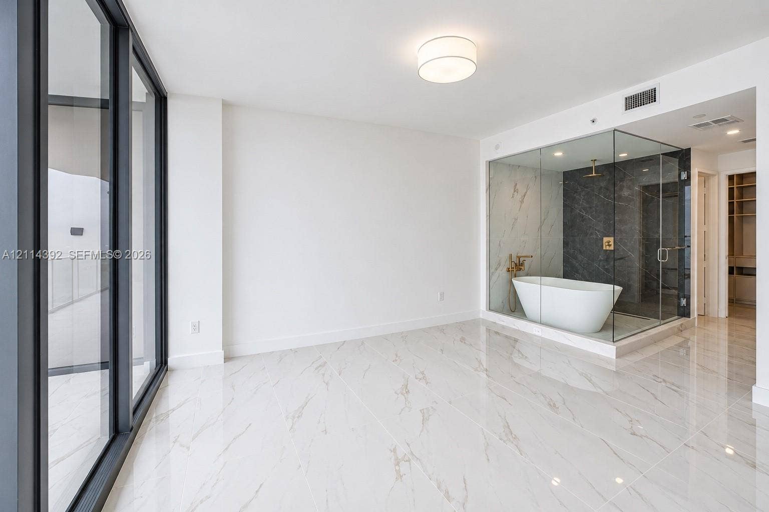 Floor-to-ceiling glass paneling, polished stone-finish flooring, glass-enclosed shower, freestanding soaking tub, and gold-tone fixtures