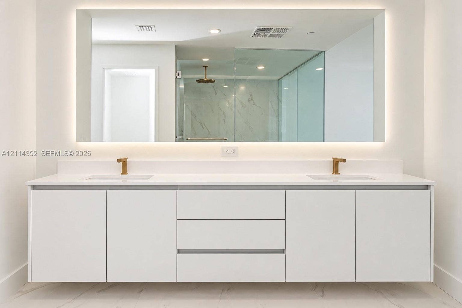 Floating white dual-sink vanity with a solid surface countertop, metallic finish faucets, a large rectangular mirror with integrated perimeter lighting, and a glass-enclosed shower with tiled wall surrounds