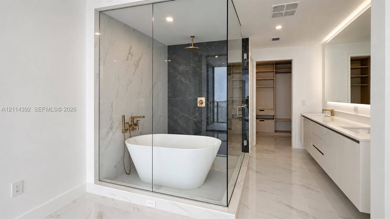 Glass-enclosed wet room featuring a freestanding soaking tub, dual-tone tile surround, and brushed gold fixtures, complemented by a dual-sink vanity with under-cabinet lighting and a walk-in dressing area with built-in shelving