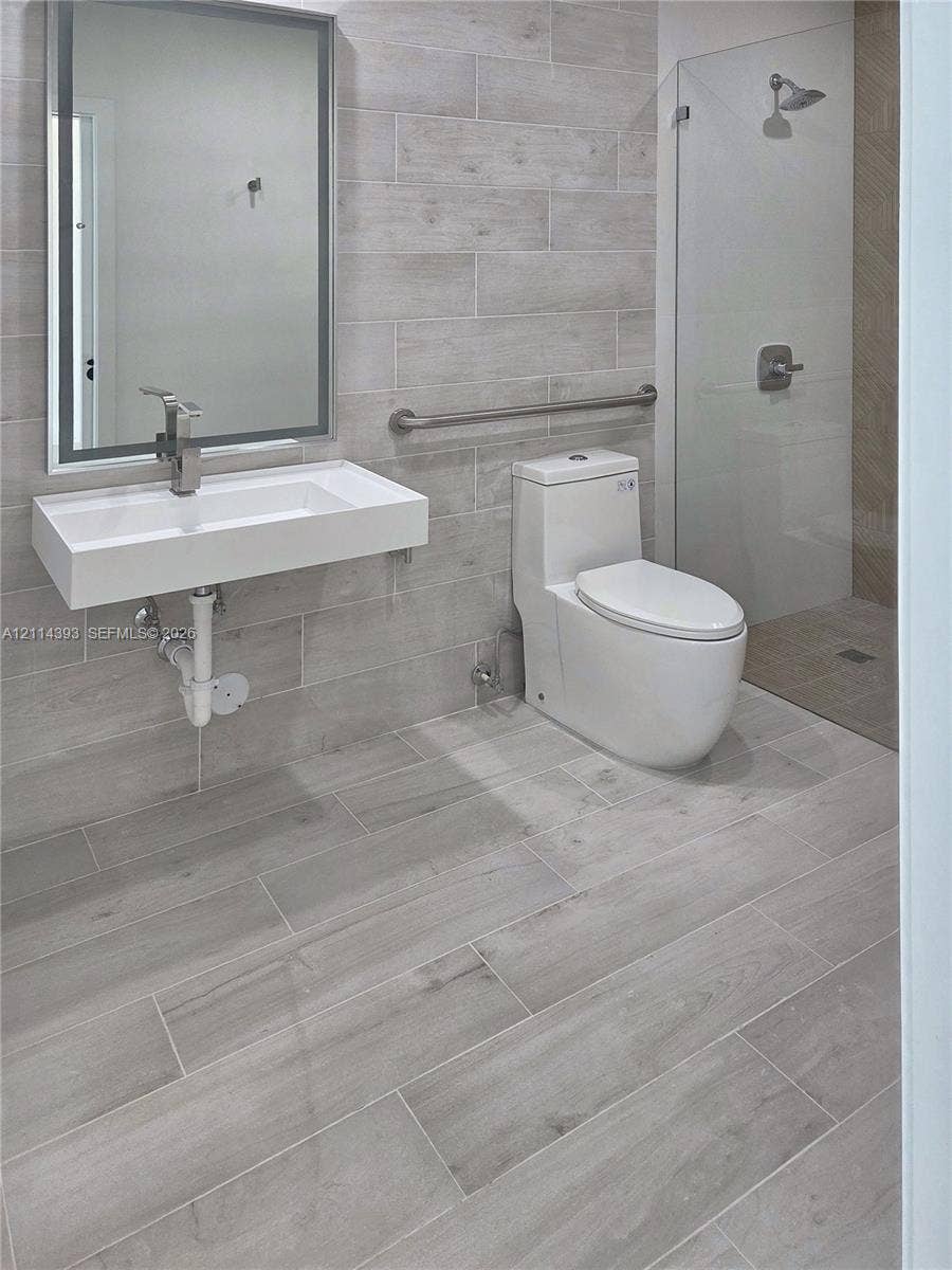 401 Pembroke Road, #202, Hallandale Beach, Hallandale Beach, FL 33009 Wall-mounted rectangular basin with a single-handle faucet, full-height tile wall surfacing, walk-in glass shower enclosure, elongated one-piece toilet, and wood-finish tile flooring