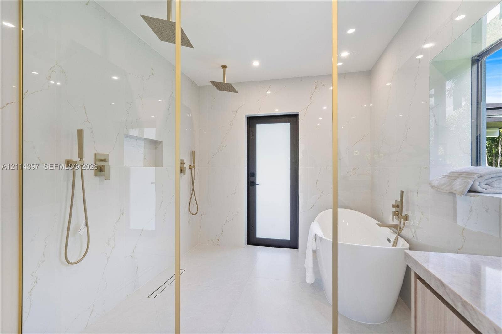 14421 SW 85th Avenue, Palmetto Bay, FL 33158 Walk-in wet room featuring oversized wall-to-wall stone-finish tile, dual ceiling-mounted rainfall showerheads, brass-finish fixtures, a freestanding soaking tub, and a built-in shower niche