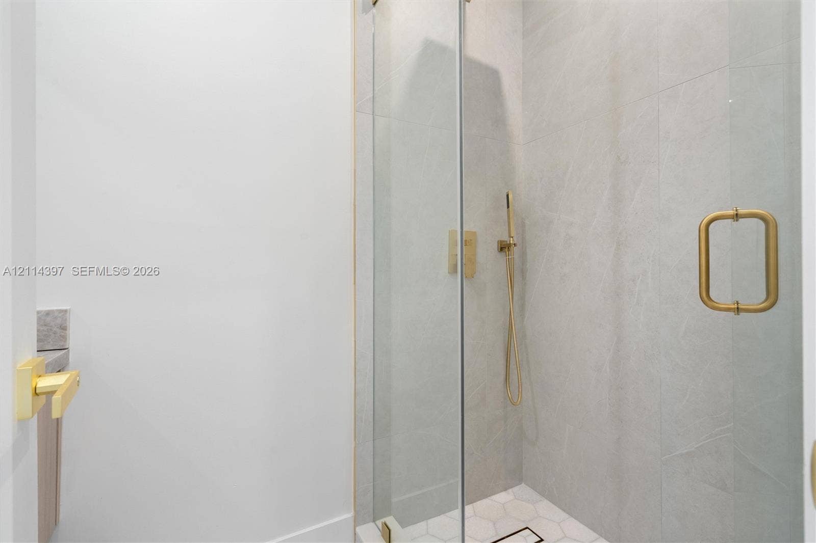 14421 SW 85th Avenue, Palmetto Bay, FL 33158 Walk-in shower featuring large-format grey tile walls, white hexagonal floor tile, brass-finish handheld faucet, and a frameless glass door with a coordinated brass handle