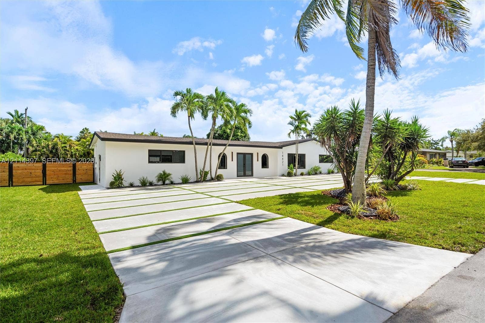 14421 SW 85th Avenue, Palmetto Bay, FL 33158 Single-story residence featuring a smooth stucco exterior, expansive concrete driveway with grass inlays, arched entryway, horizontal wood-slat privacy fencing, and mature palm landscaping