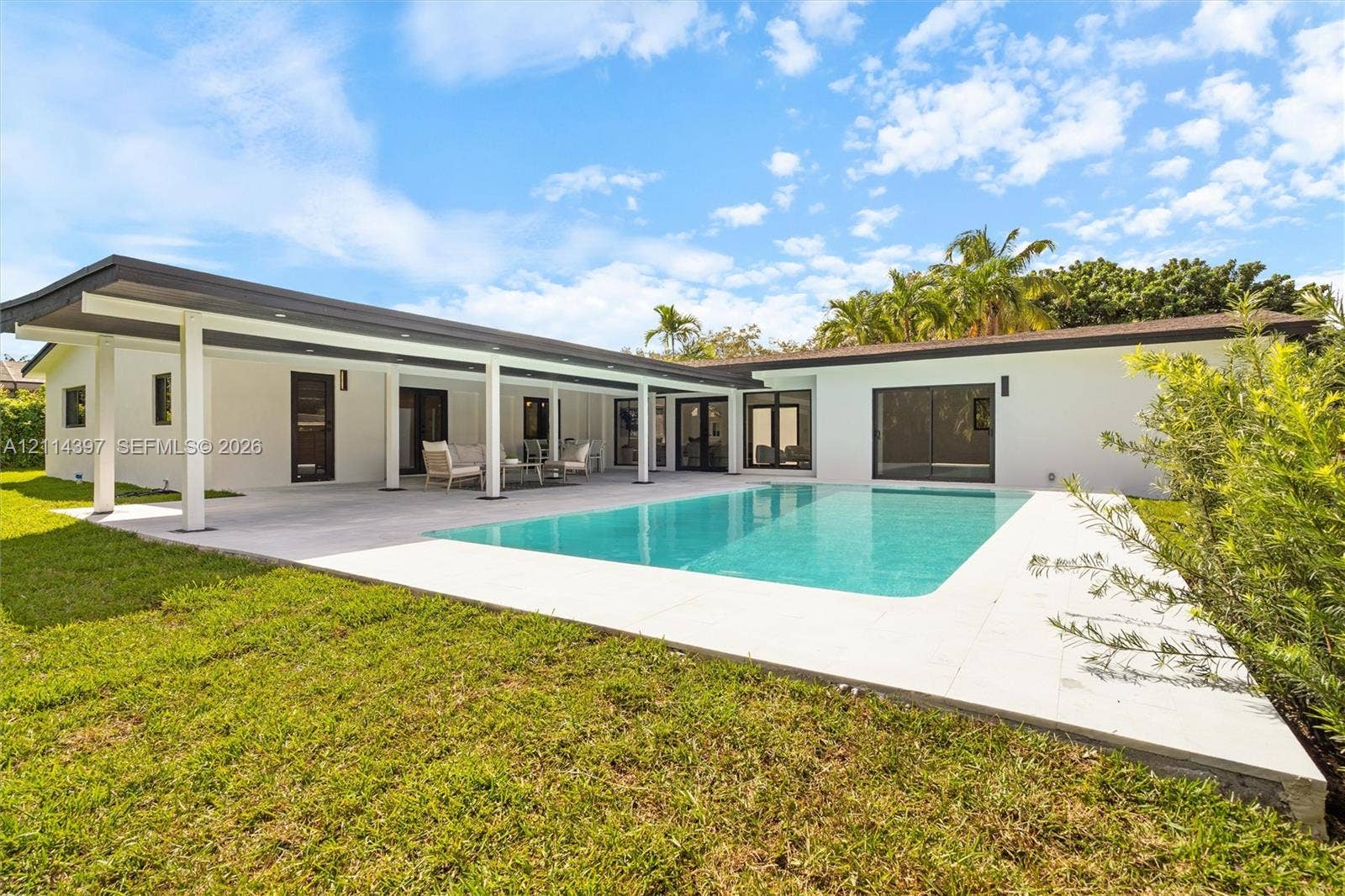 14421 SW 85th Avenue, Palmetto Bay, FL 33158 Exterior featuring a spacious covered patio, a rectangular in-ground swimming pool, light-toned stone pool decking, and white stucco siding