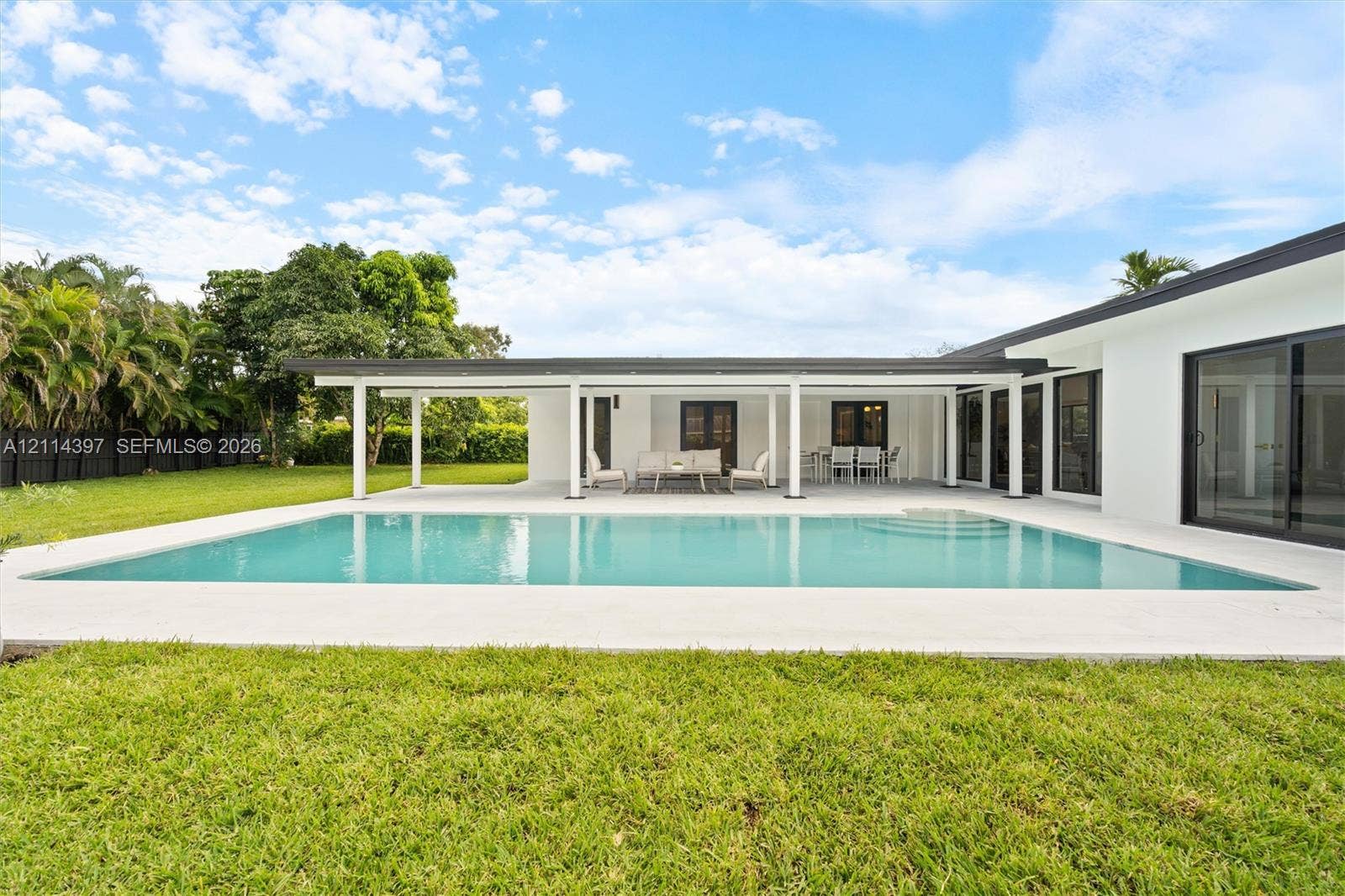 14421 SW 85th Avenue, Palmetto Bay, FL 33158 Backyard featuring a rectangular swimming pool, expansive concrete patio, covered lanai with structural pillars, sliding glass door panels, and manicured lawn