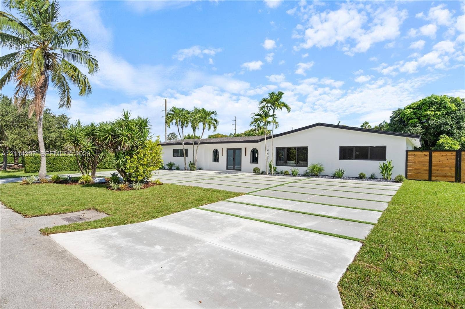 14421 SW 85th Avenue, Palmetto Bay, FL 33158 Single-story residence featuring a smooth white exterior, expansive concrete driveway with inset grass turf strips, mature palm trees, and horizontal wood-panel fencing