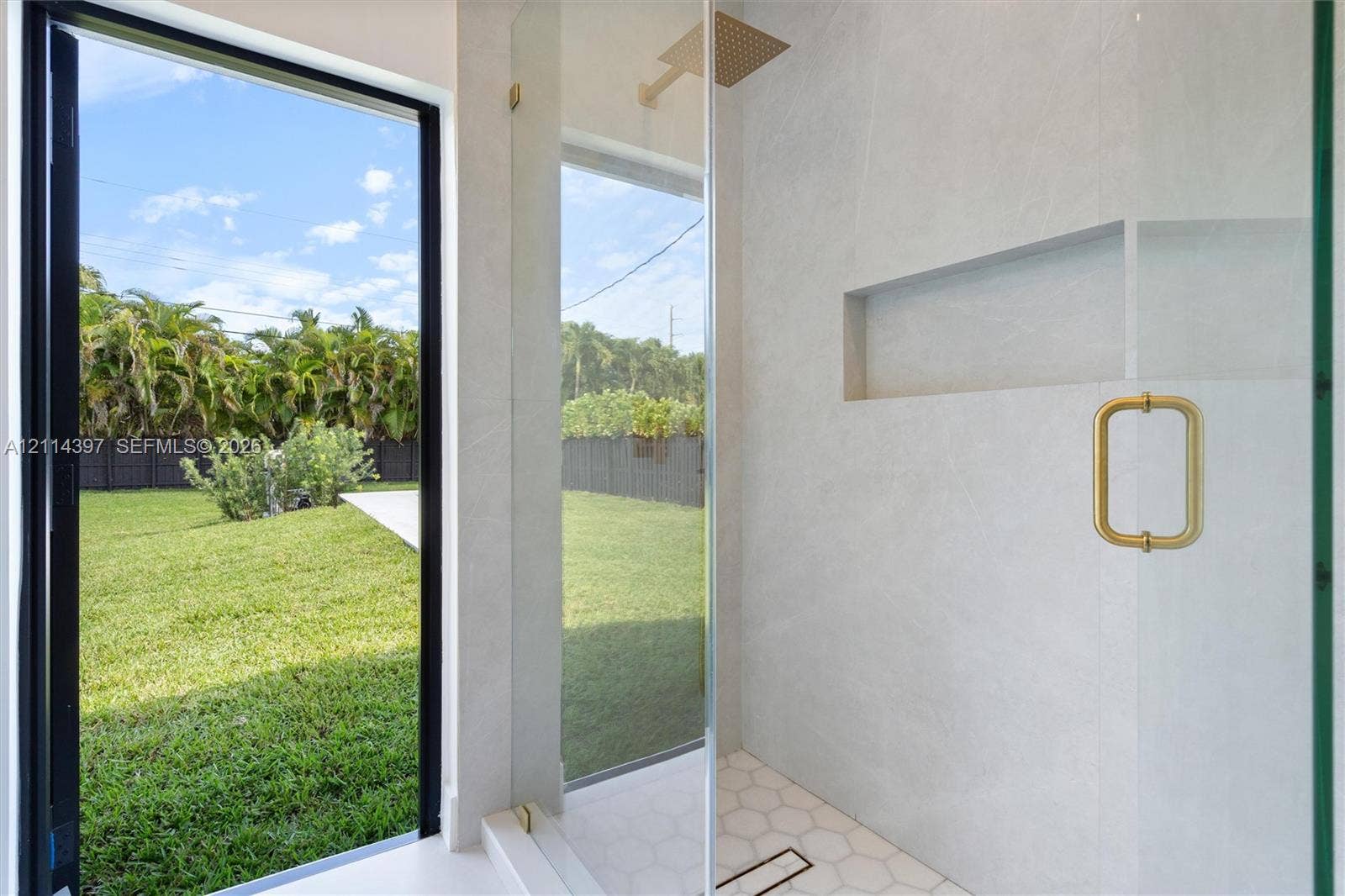 14421 SW 85th Avenue, Palmetto Bay, FL 33158 Walk-in shower featuring large-format stone-tile walls, hexagonal floor tiling, a recessed wall niche, a brass-finish rainfall showerhead, and a floor-to-ceiling glass panel with a matching brass-finish handle