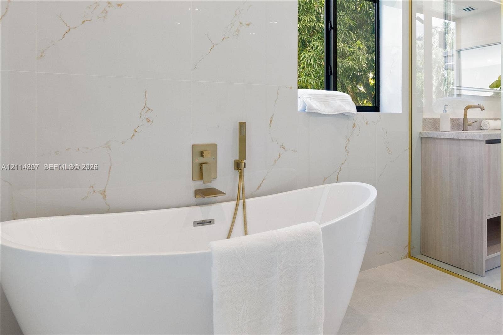 14421 SW 85th Avenue, Palmetto Bay, FL 33158 Freestanding soaking tub featuring wall-mounted metallic fixtures and a handheld shower spray, set against large-format tile walls with prominent veining