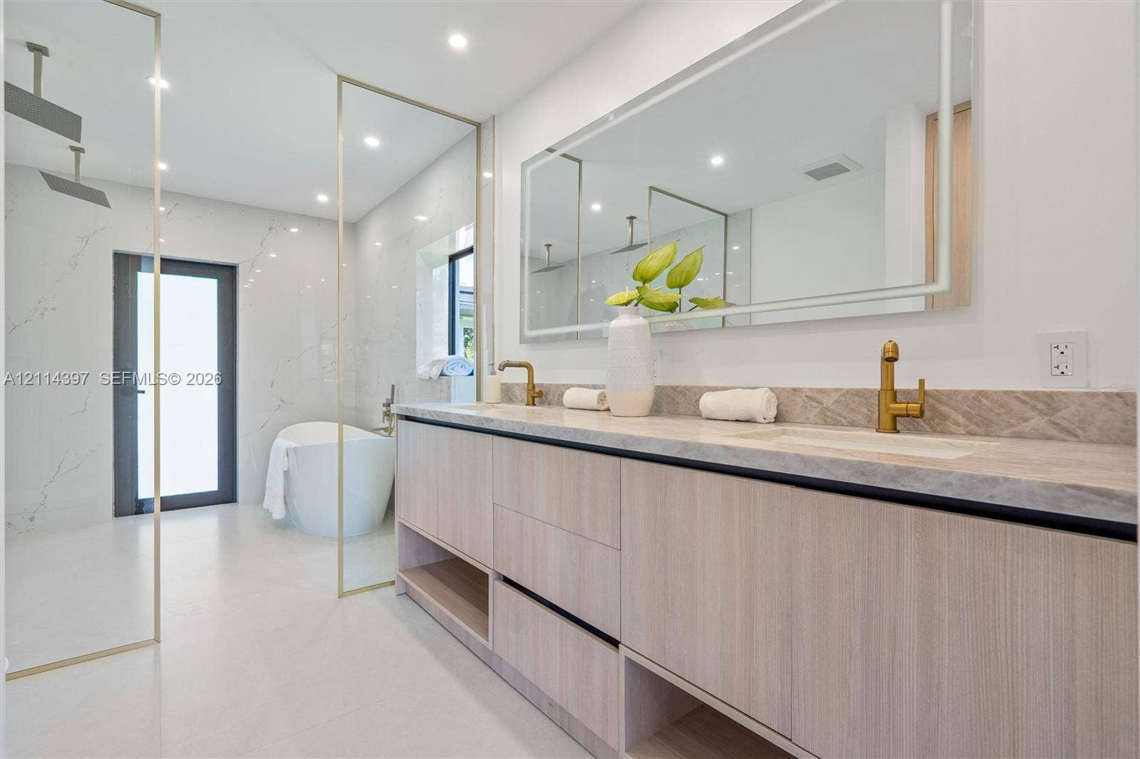 14421 SW 85th Avenue, Palmetto Bay, FL 33158 Dual-sink vanity with wood-finish cabinetry, natural stone countertop, brass fixtures, integrated LED-backlit mirror, and glass-enclosed wet room featuring a soaking tub
