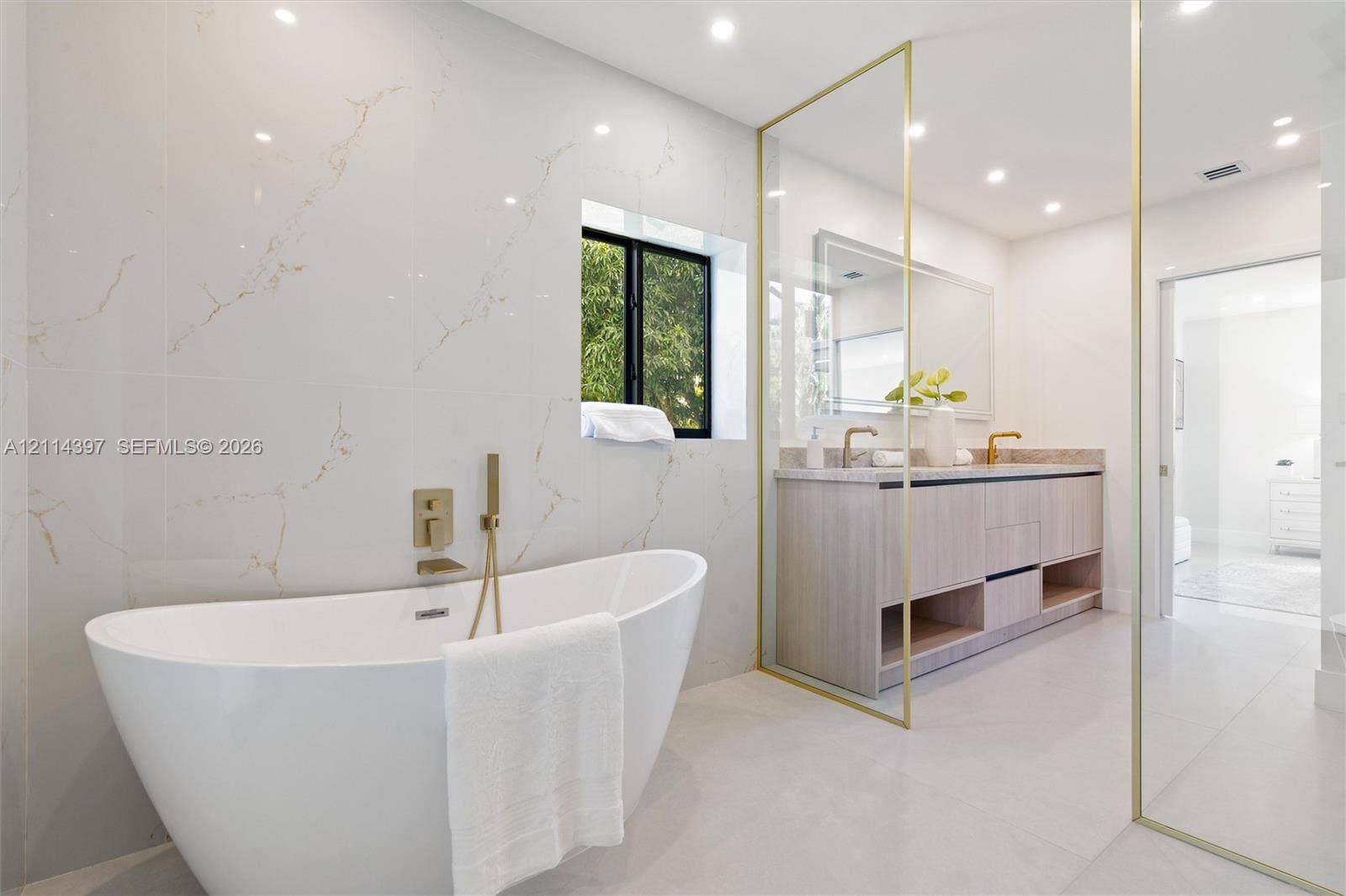 14421 SW 85th Avenue, Palmetto Bay, FL 33158 Freestanding soaking tub with gold-tone wall-mounted faucet, large-format tile wall cladding, wood-finish floating vanity with dual sinks, gold-trimmed glass enclosure, and recessed lighting