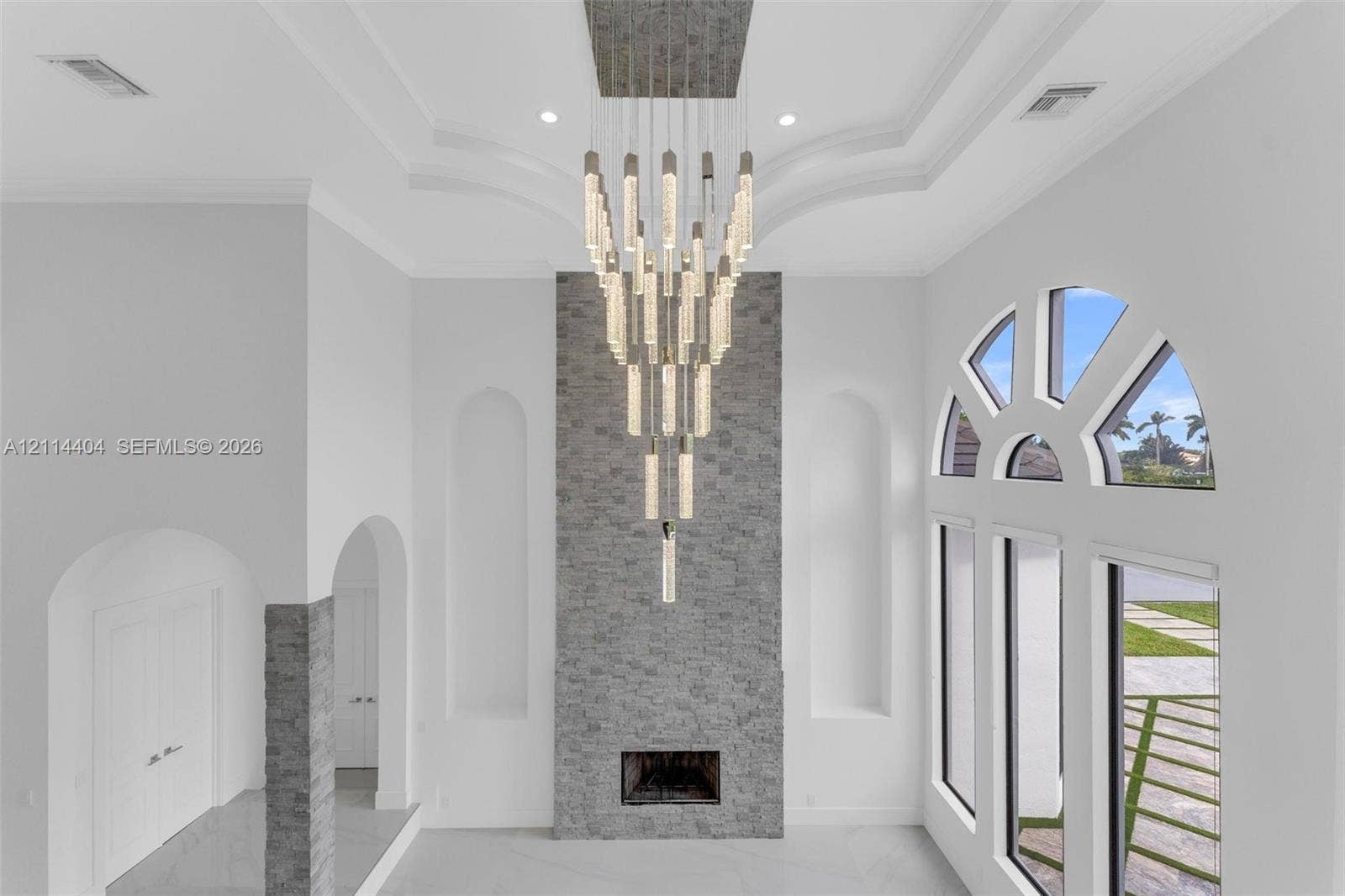 High-ceilinged interior featuring a tiered crystal chandelier, a floor-to-ceiling stone fireplace surround, dual arched wall niches, and a multi-pane arched window