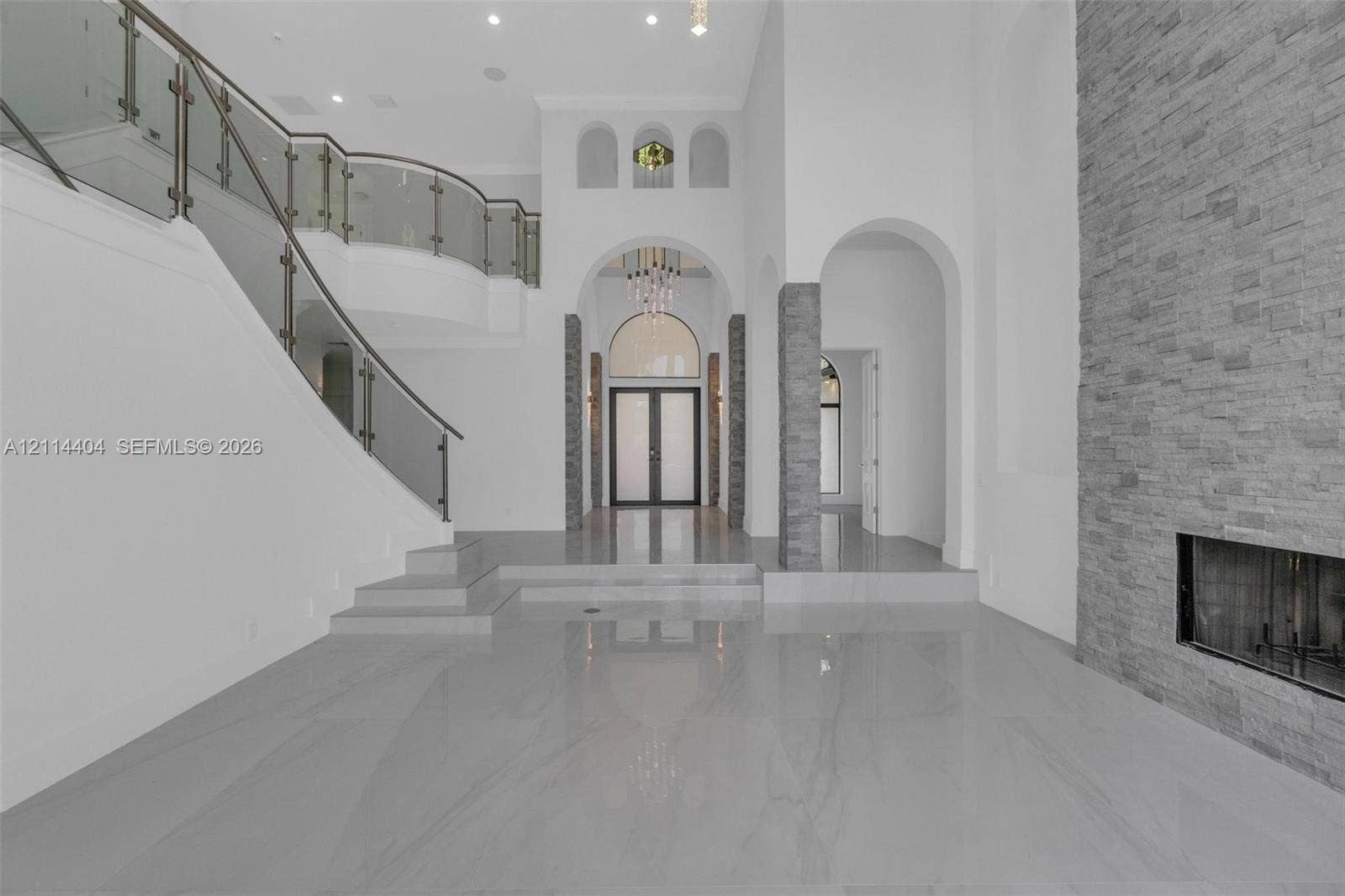 Grand foyer featuring polished stone flooring, a sweeping staircase with glass railings, soaring arched entryways, and a floor-to-ceiling stacked stone fireplace