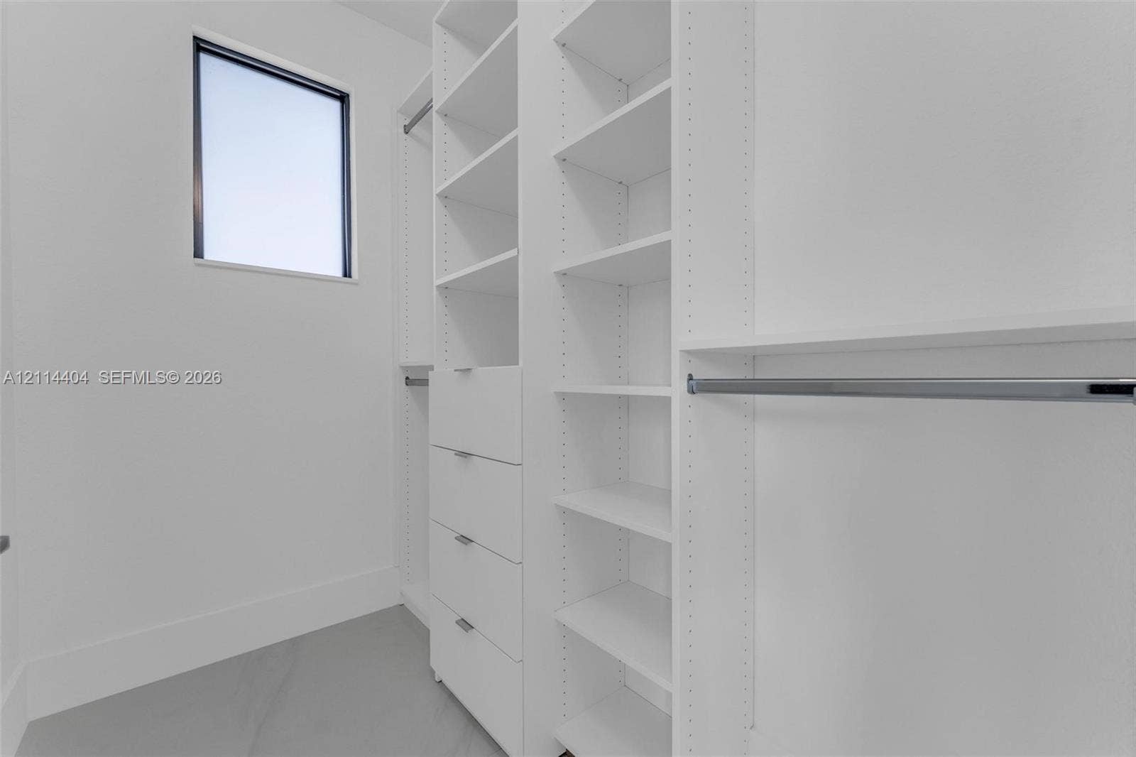 Custom walk-in closet featuring white built-in shelving, a four-drawer dresser unit, metallic clothing rods, and a rectangular window with a dark frame