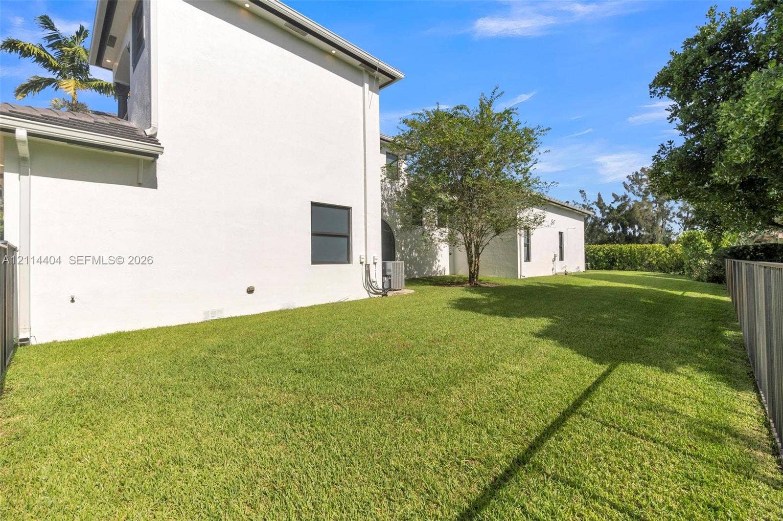 Expansive rear yard featuring a manicured lawn, perimeter fencing, mature trees, and light-toned stucco exterior siding