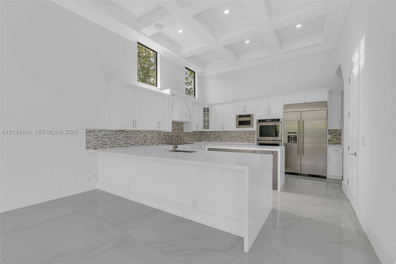Kitchen featuring a coffered ceiling, waterfall-edge quartz island, stainless steel appliances, stacked-stone backsplash, and cabinetry with integrated recessed lighting