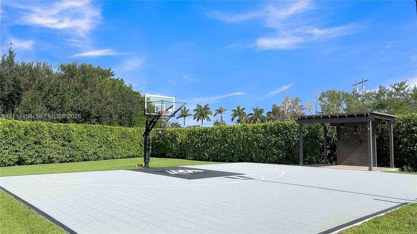 Outdoor athletic court featuring a multi-sport tile surface, built-in basketball hoop, and a covered pavilion structure, all enclosed by mature perimeter landscaping