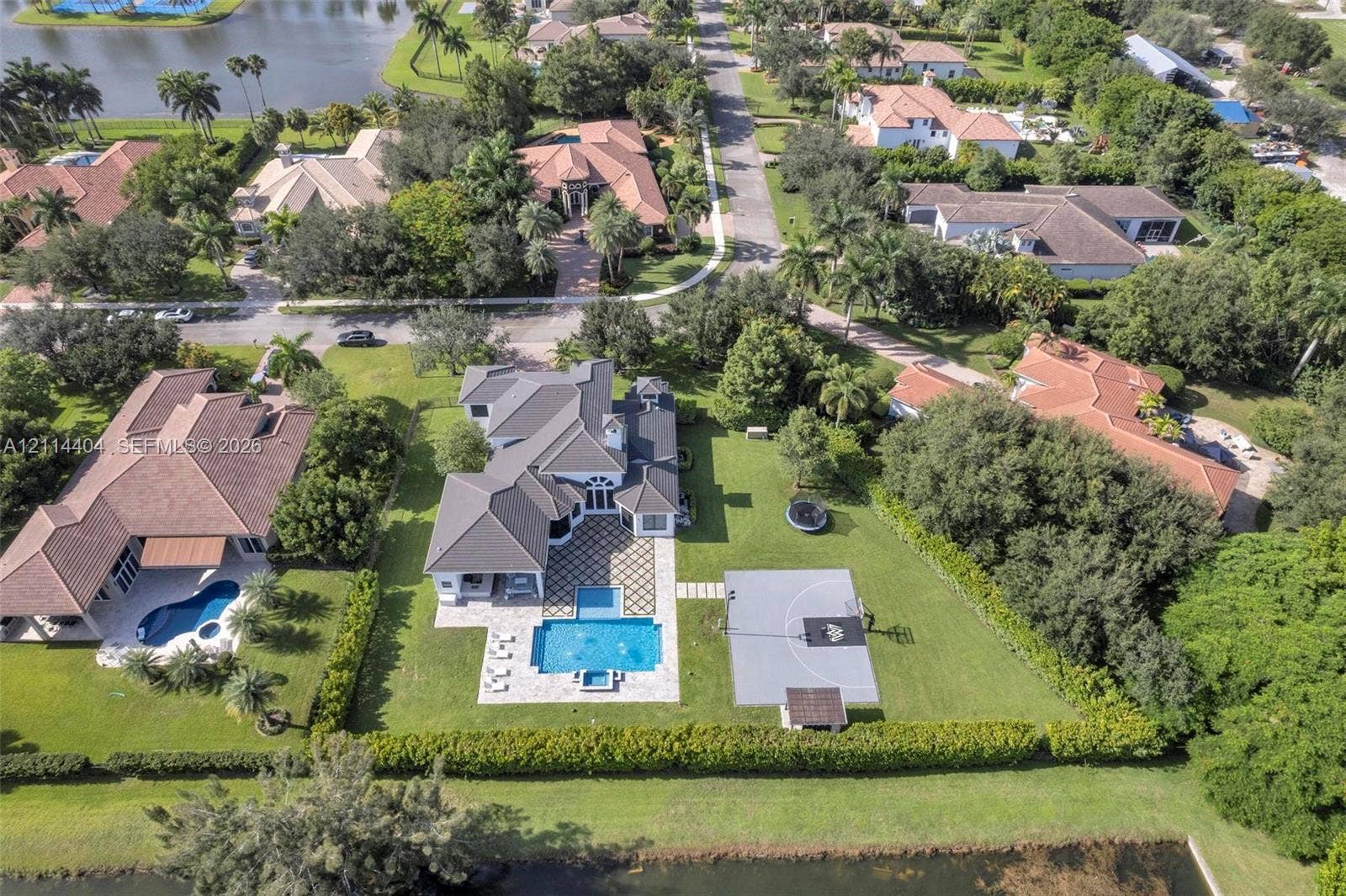 Aerial view featuring a multi-gabled residence with a paved patio, a rectangular swimming pool, and a concrete sports court set on a landscaped lot with perimeter privacy hedging