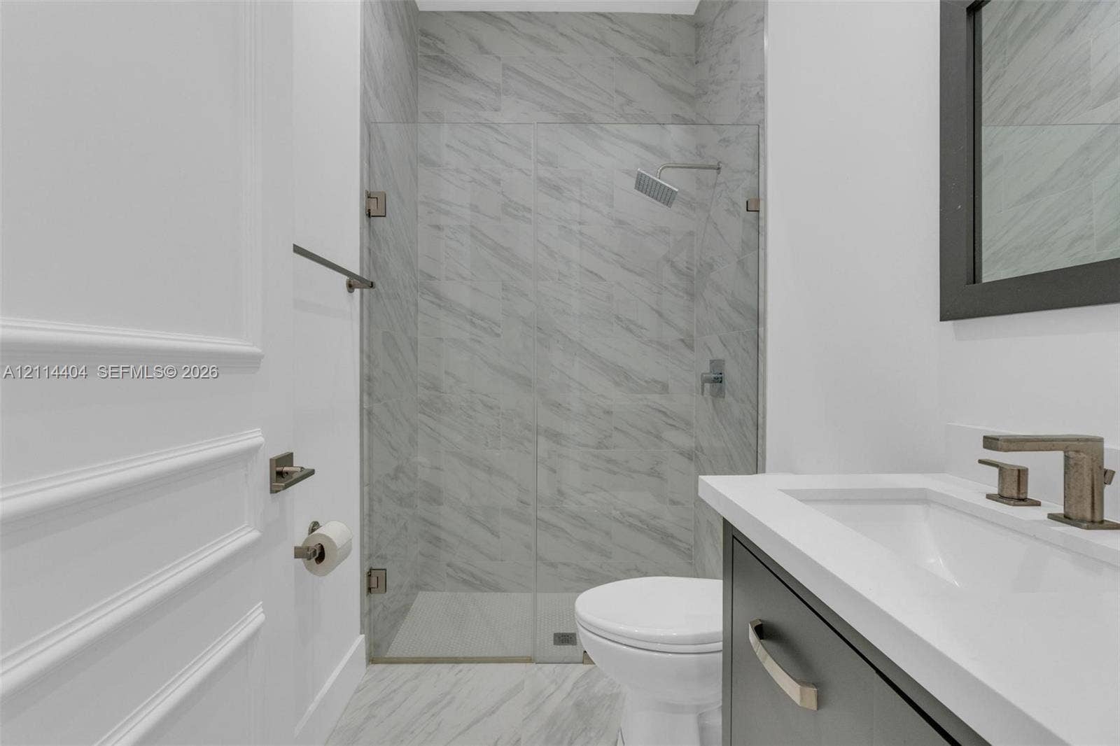 Bathroom featuring a walk-in shower with stone-patterned tile, frameless glass enclosure, wall-mounted rainfall showerhead, and a vanity with a white countertop and dark cabinetry