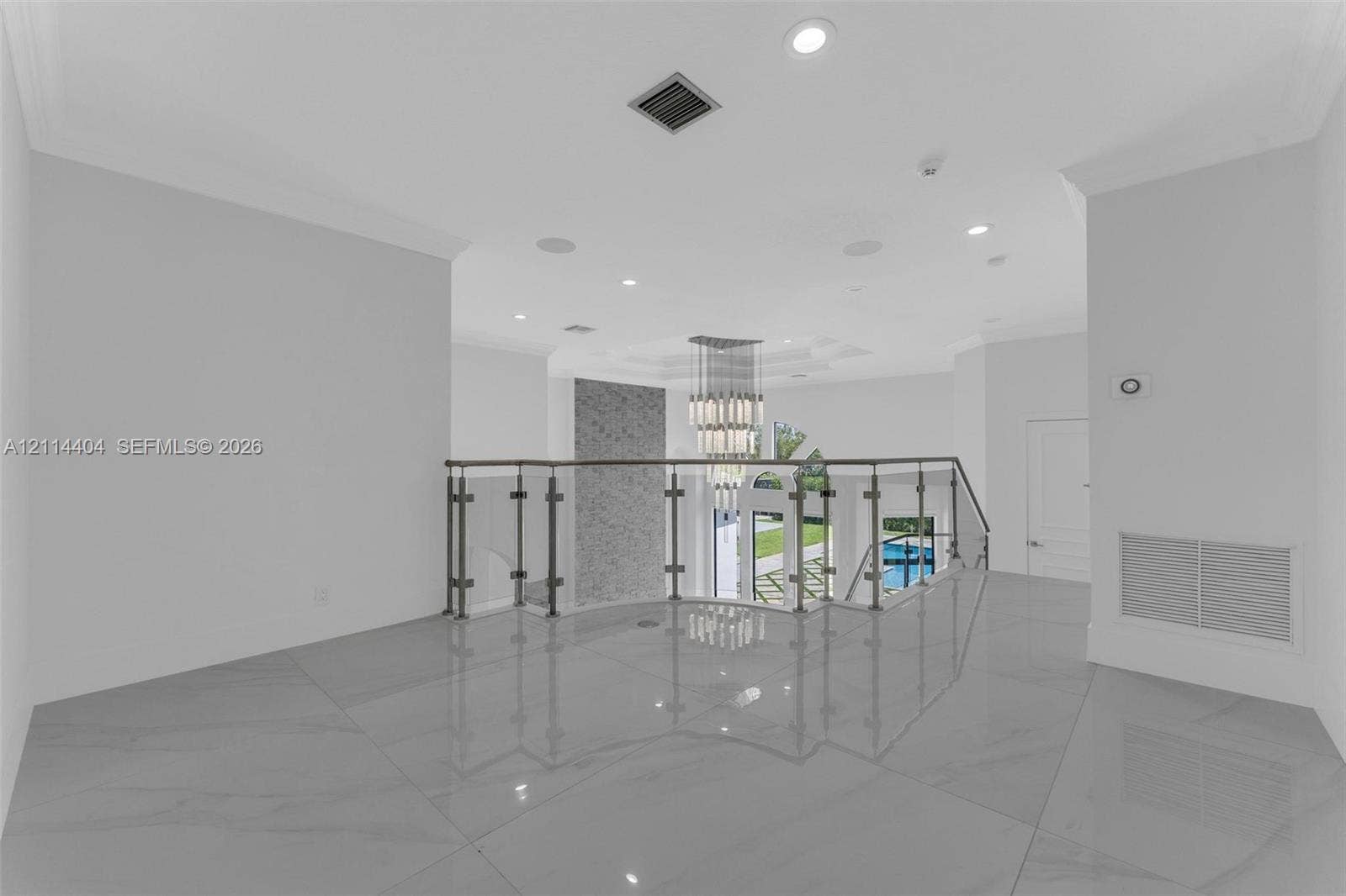 Open landing featuring large-format tile flooring, a tiered glass-and-metal chandelier, glass-panel railing with metal posts, and a textured stone-finish accent wall