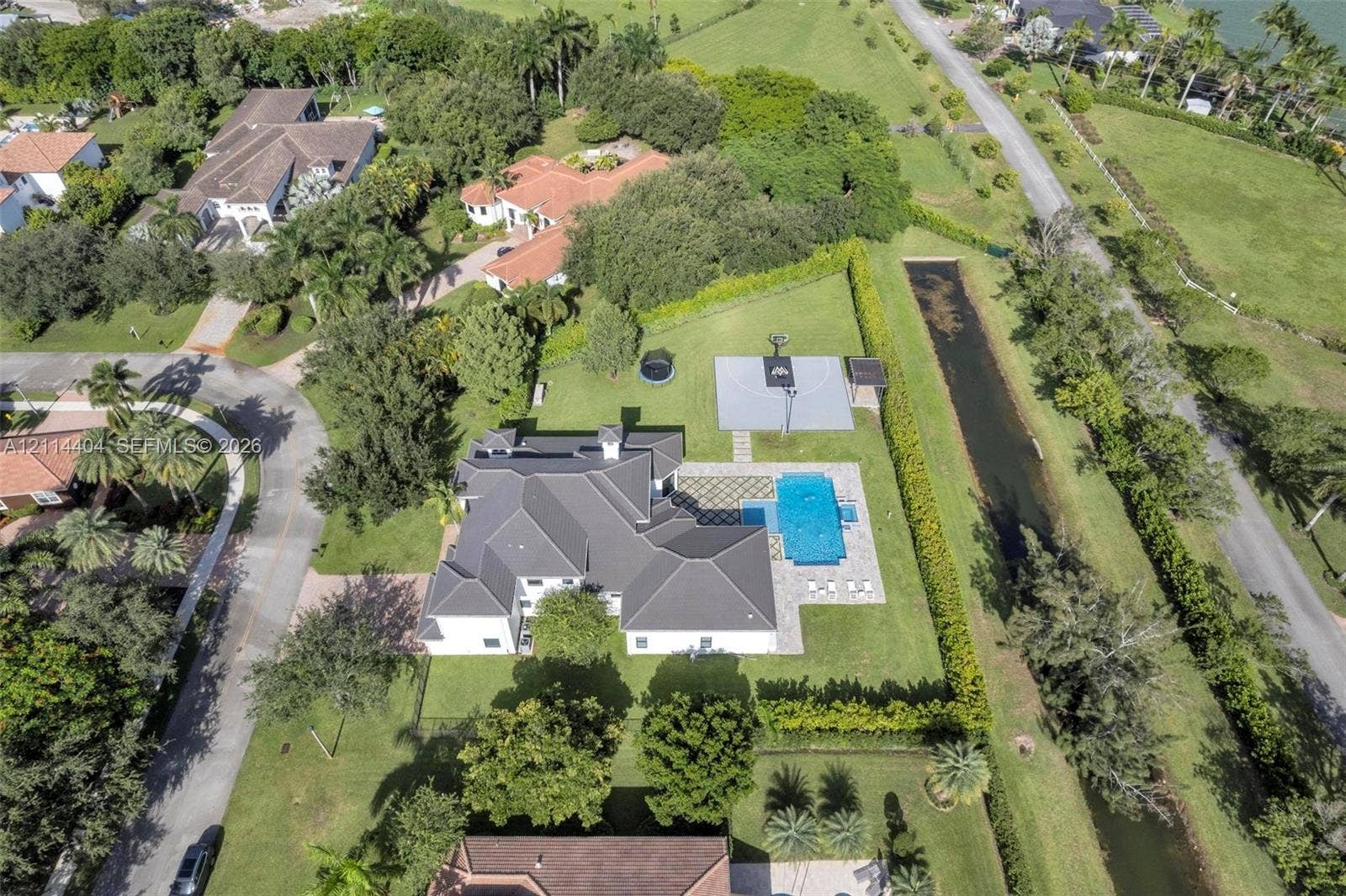 Aerial perspective featuring a multi-gabled roofline, a private swimming pool with patterned stone decking, a paved sports court, and mature landscaping