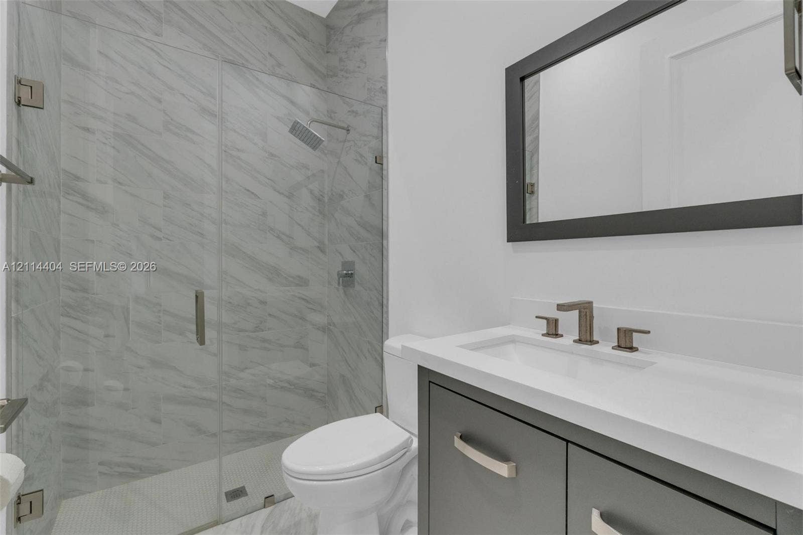 Modern vanity with a rectangular sink, stone-finish countertop, and dark-toned cabinetry, paired with a glass-enclosed shower featuring large-format tile surrounds, a rain-style showerhead, and a framed mirror