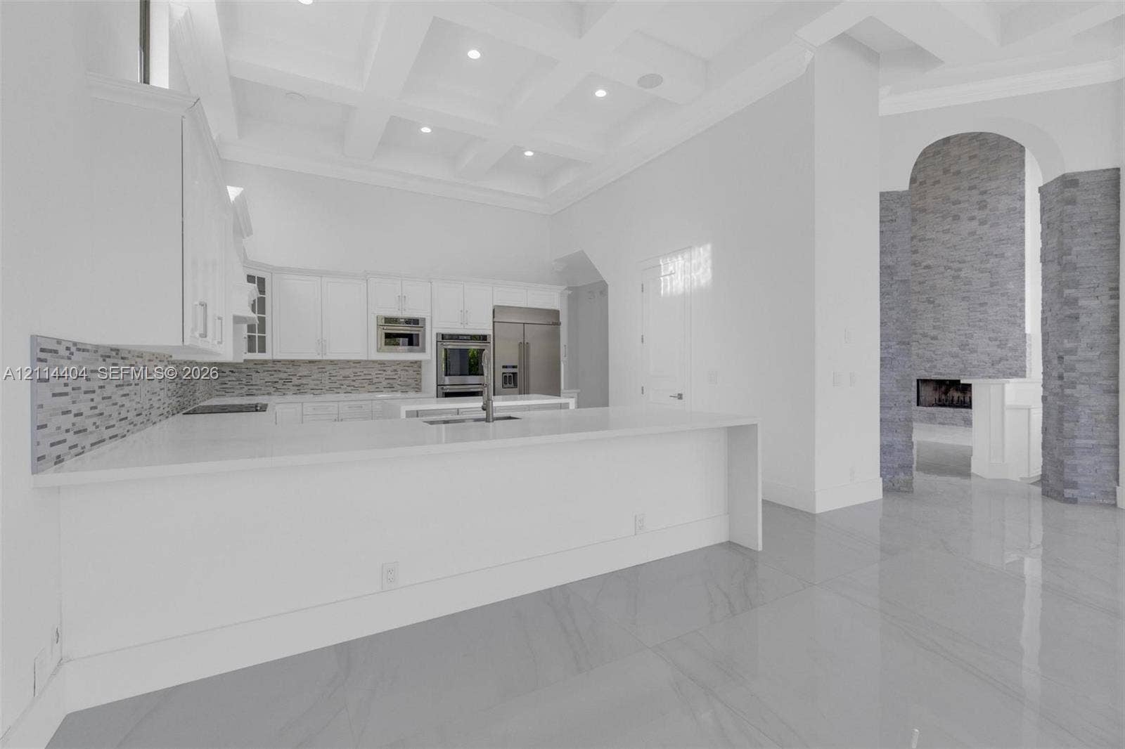 Kitchen featuring custom cabinetry, a stone-tile backsplash, expansive solid-surface island, and coffered ceilings, complemented by a floor-to-ceiling stone fireplace surround