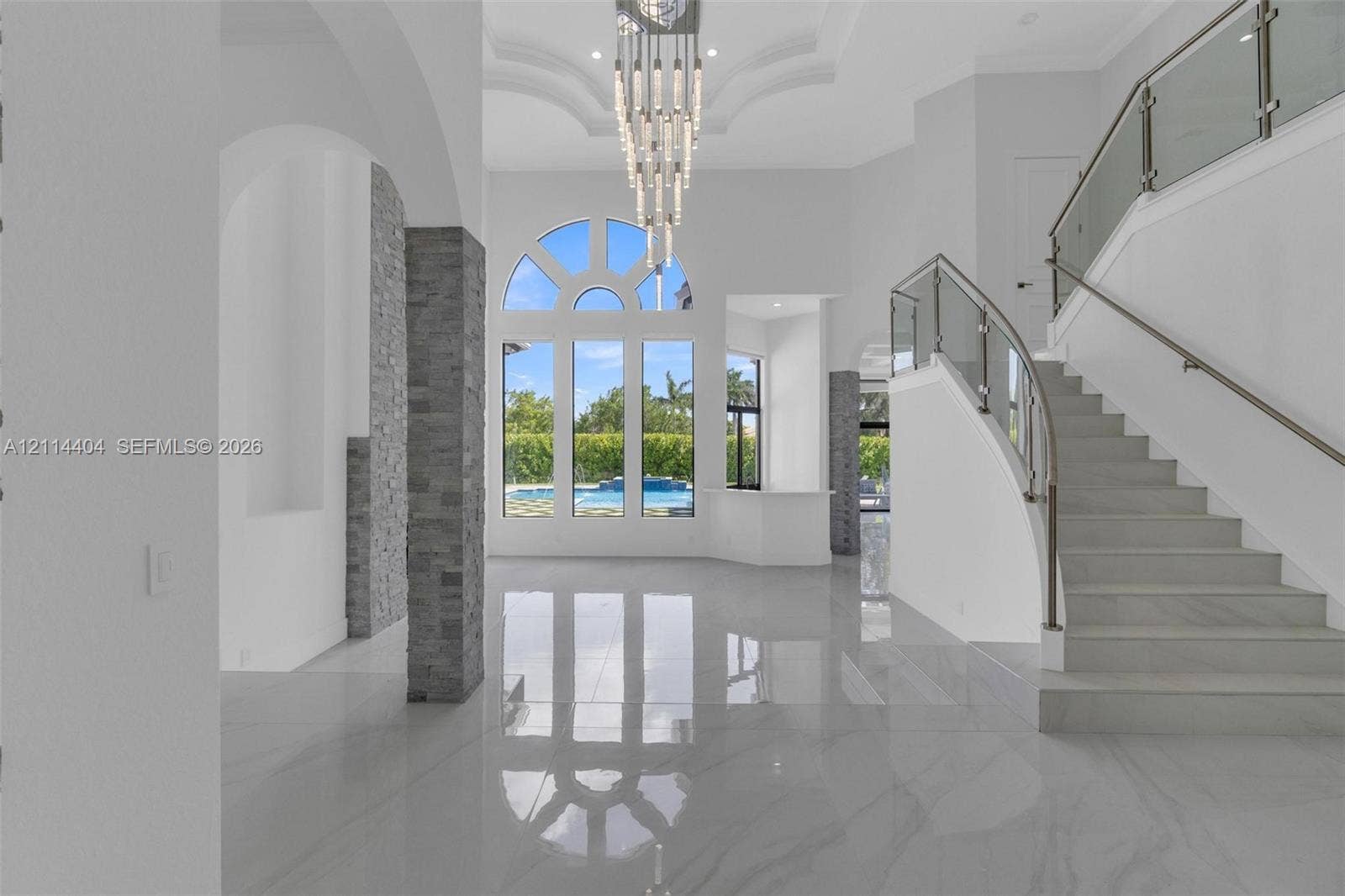 Grand foyer featuring polished stone flooring, a multi-tiered crystal chandelier, vaulted ceilings with recessed molding, a curved staircase with glass railings, and floor-to-ceiling arched windows with stacked stone pillars