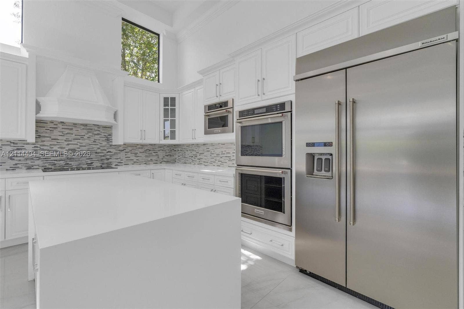 Kitchen featuring white cabinetry, an expansive central island with a solid-surface countertop, built-in stainless steel double ovens, a full-size stainless steel refrigerator, and a mosaic tile backsplash