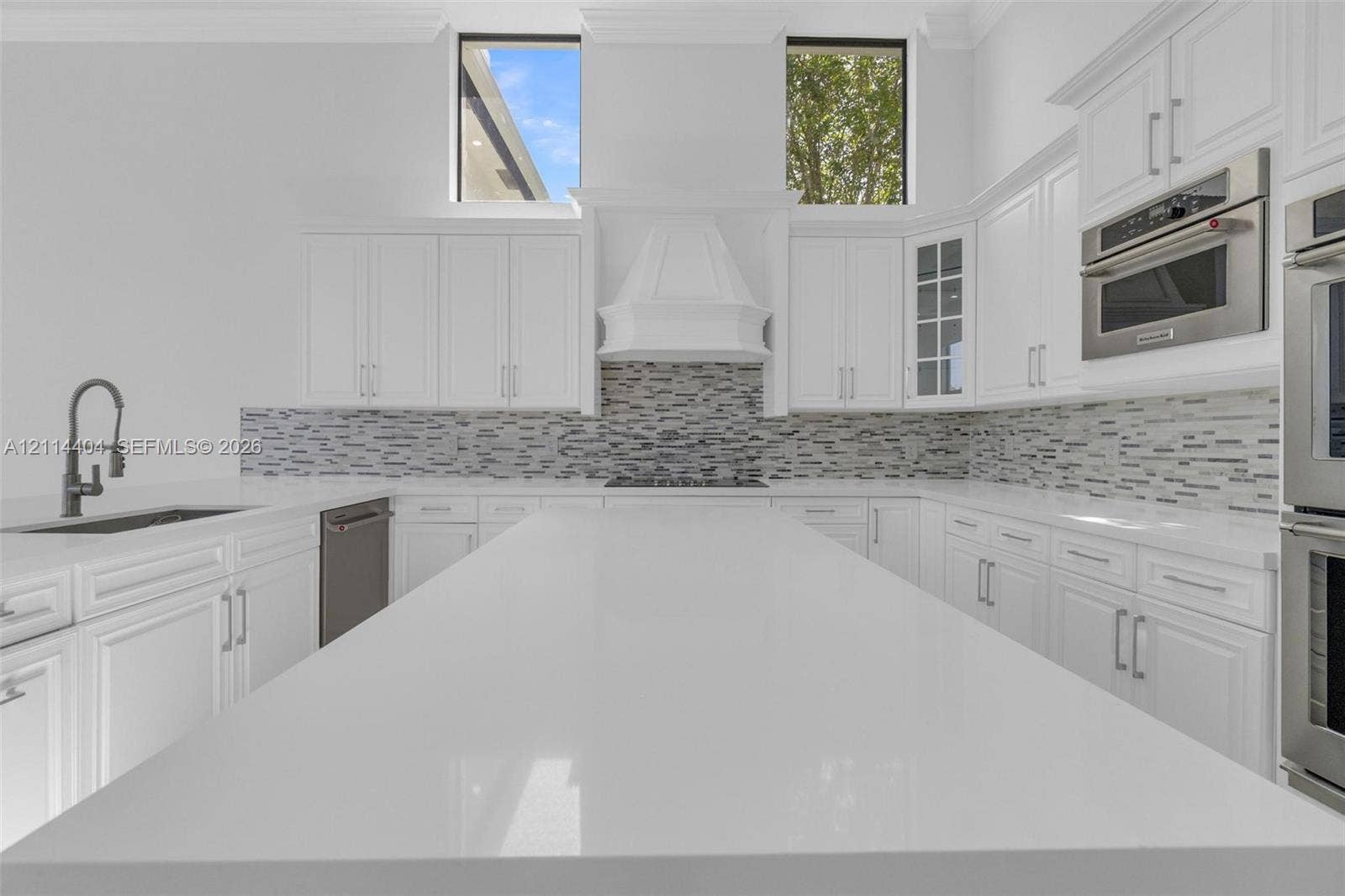 Kitchen featuring a large central island with solid-surface countertop, white shaker-style cabinetry, a multi-tonal linear tile backsplash, high-ceiling transoms, and built-in stainless steel appliances
