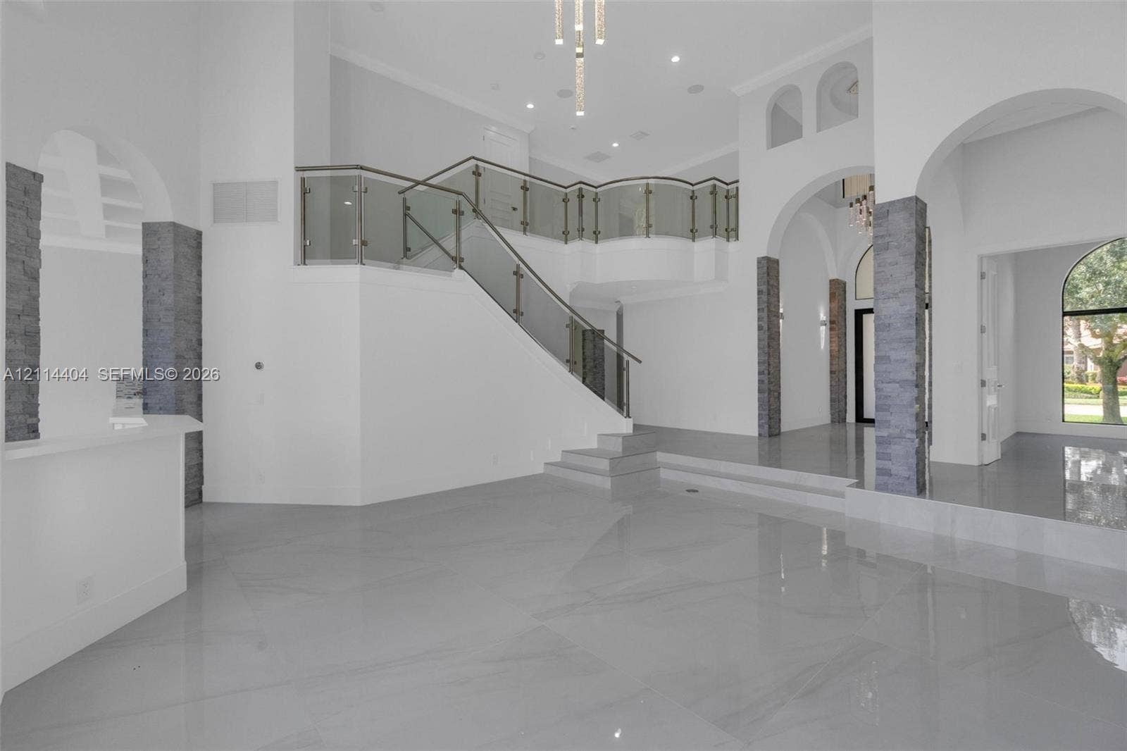 Grand foyer featuring large-format tile flooring, tiered levels, architectural stone-veneer pillars, glass-panel railings, and arched entryways