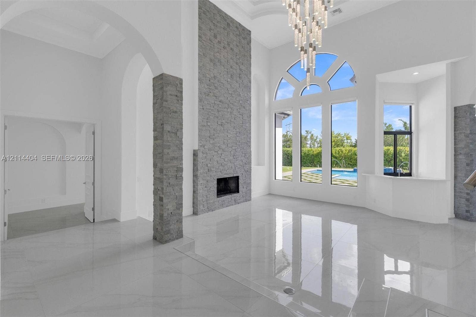 Double-height ceilings featuring an expansive stacked-stone fireplace, polished tile flooring, arched architectural detailing, a multi-tiered crystal chandelier, and large-scale windows with exterior pool exposure