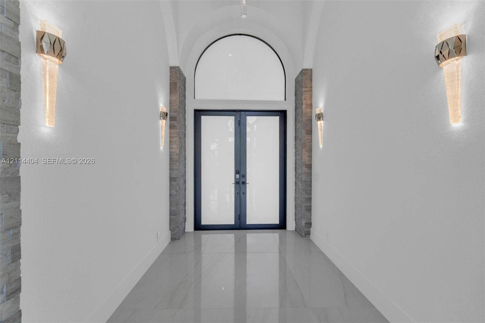 Entryway featuring arched architectural detailing, double glass-panel doors, stone-finish columns, and large-format tile flooring