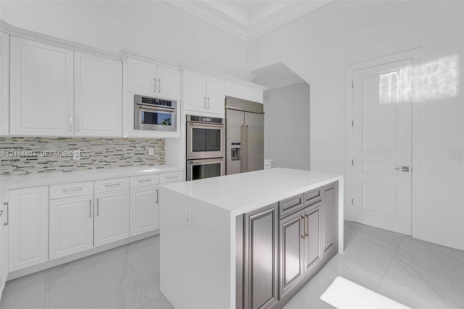 Kitchen featuring white shaker cabinetry, stone-surface countertops, a contrasting grey center island, stainless steel wall-mounted double ovens, and a multi-toned linear backsplash