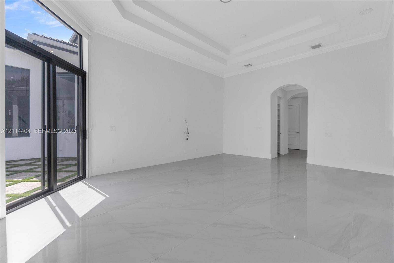 High-ceiling interior featuring a multi-tiered tray ceiling, floor-to-ceiling glass sliding doors, polished stone-finish flooring, and an arched entryway