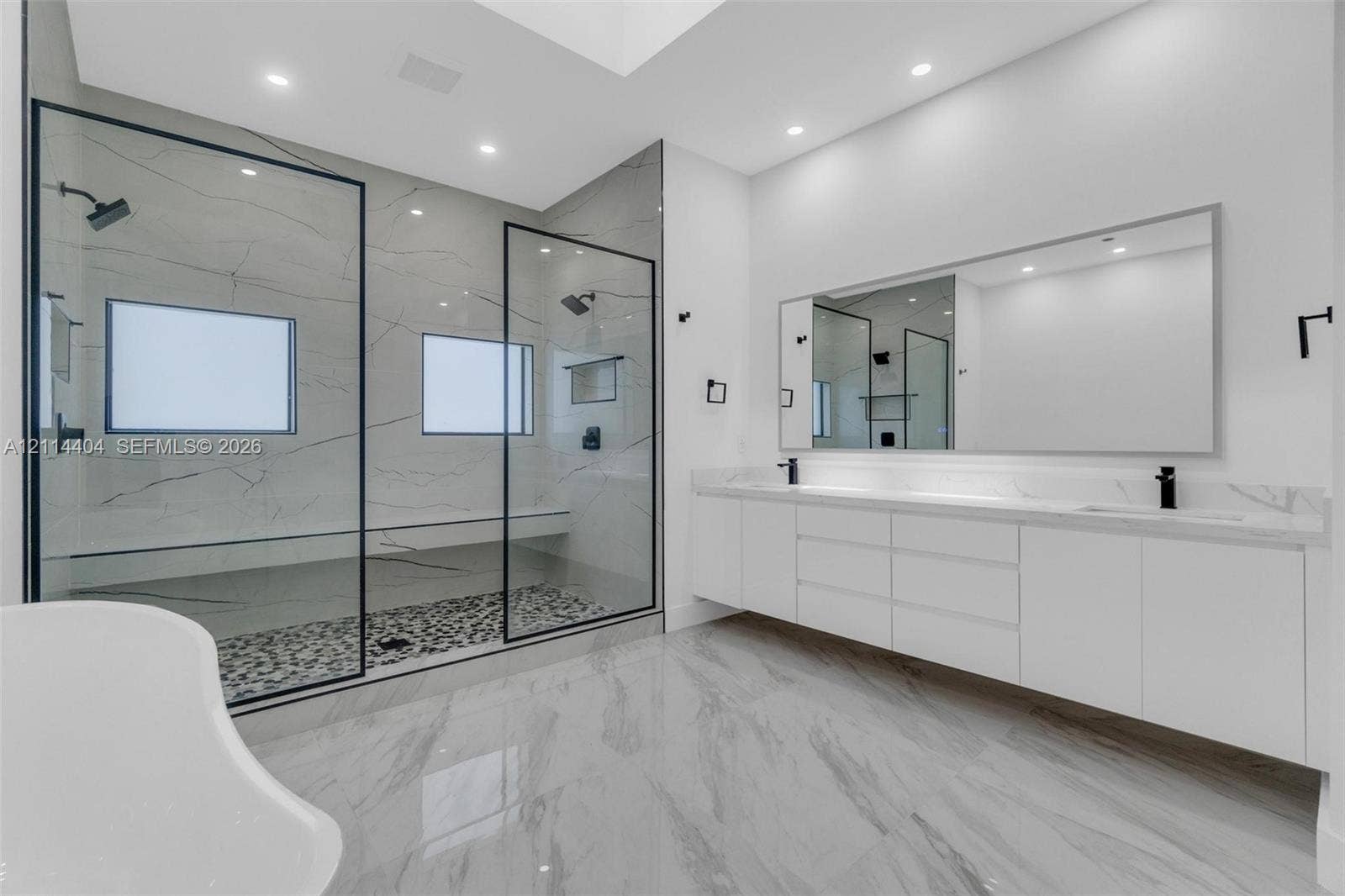 Double vanity with integrated sinks, expansive walk-in shower featuring a stone-tile floor and matte black hardware, large frameless mirror, and recessed ceiling lighting