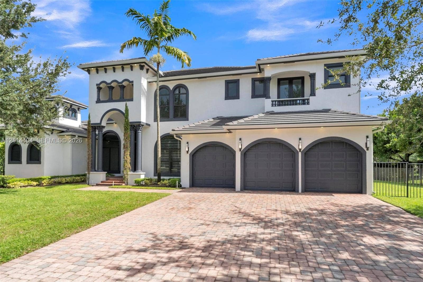 Mediterranean-style exterior featuring smooth stucco finish, arched entryway with decorative columns, three-car garage with arched doors, second-level balcony, and brick-paver driveway
