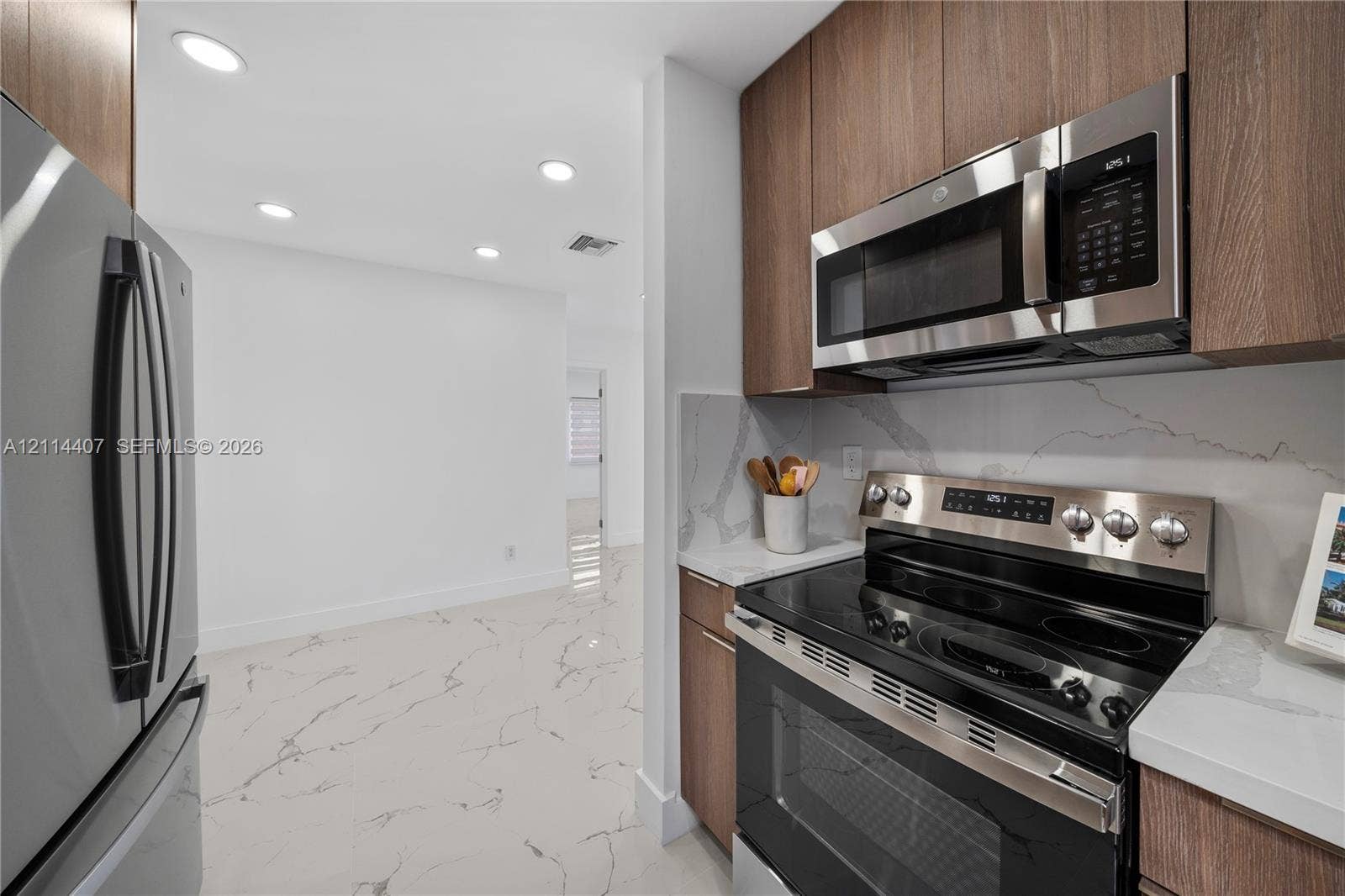 Kitchen featuring wood-finish cabinetry, stainless steel appliances, stone-patterned countertops, matching stone-patterned backsplash, and large-format tile flooring