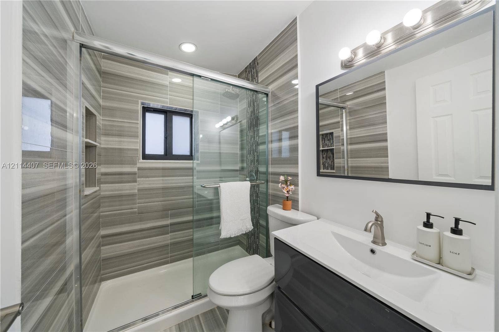 Bathroom featuring a glass-enclosed walk-in shower with horizontal-striped tile, floating vanity with a white countertop and integrated sink, and a rectangular mirror with black frame