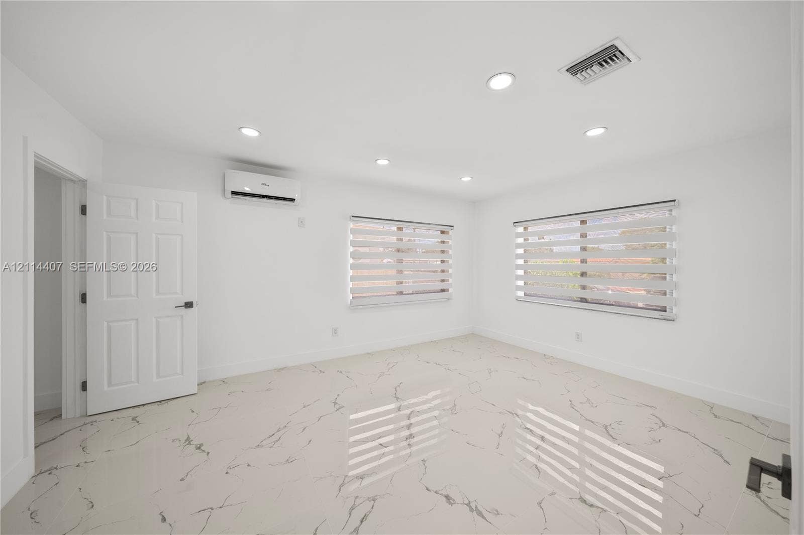 Large-format tile flooring with a neutral veined pattern, recessed ceiling lighting, wall-mounted ductless climate control unit, and dual-layered cellular shade window coverings