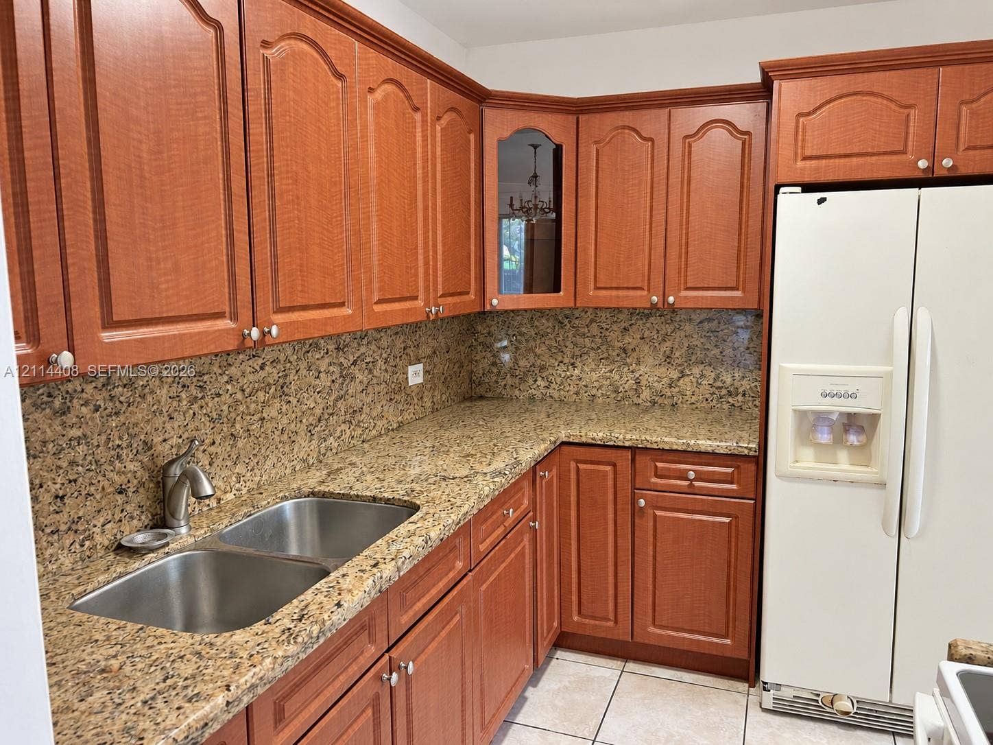 Kitchen featuring wood cabinetry with arched detailing, natural stone countertops, a full-height stone backsplash, a stainless steel double-basin undermount sink, and a glass-front display cabinet
